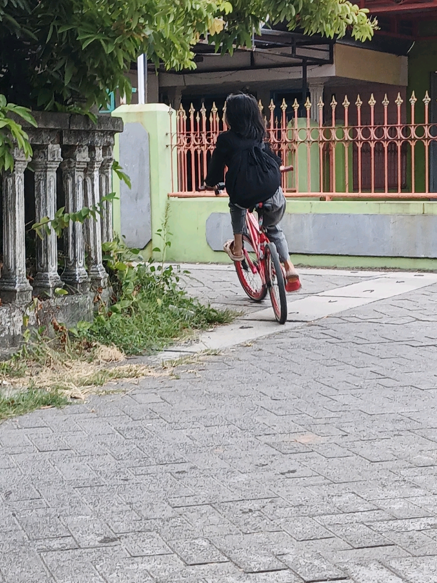 A person with long dark hair, wearing a black jacket and gray pants, is riding a red bicycle with white accents on a paved walkway. The bicycle has red wheels. The person's left foot is visible, wearing a light-colored sandal, and their right foot is also visible, wearing a red sandal. They are pedaling the bike, facing away from the camera and slightly to the left.
The walkway is made of gray interlocking pavers. To the left of the walkway is a grey stone fence with decorative pillars and some overgrown green vegetation. In the background, there is a light green wall and a decorative orange and gold metal fence. Behind the metal fence, a building with a yellow and green exterior is visible, with a red roof structure. Lush green leaves and branches from a tree hang over the scene from the top left. The overall environment suggests an outdoor setting, possibly a residential area in Ketegan, Indonesia. The lighting indicates daytime.