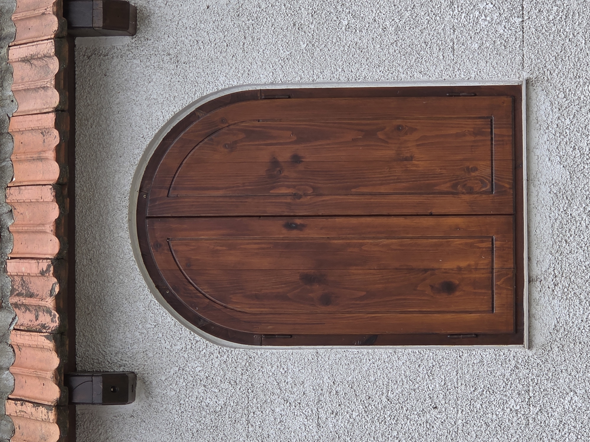 A horizontally oriented, medium-angle shot presents a section of an exterior wall in Varna, Bulgaria. The wall's surface is textured with a light gray stucco finish. In the center of the frame is a window shutter, constructed of two horizontal wooden planks finished in a dark reddish-brown stain. The shutter is rectangular in shape, with the left side rounded into a half-circle. A light gray frame surrounds the shutter and is slightly recessed into the wall. Along the left edge of the frame, terracotta-colored roof tiles are visible, stacked in a vertical column and set in mortar, creating a decorative element along the wall. Two dark brown, wooden blocks are attached to the wall to the right of the terracotta roof tiles.