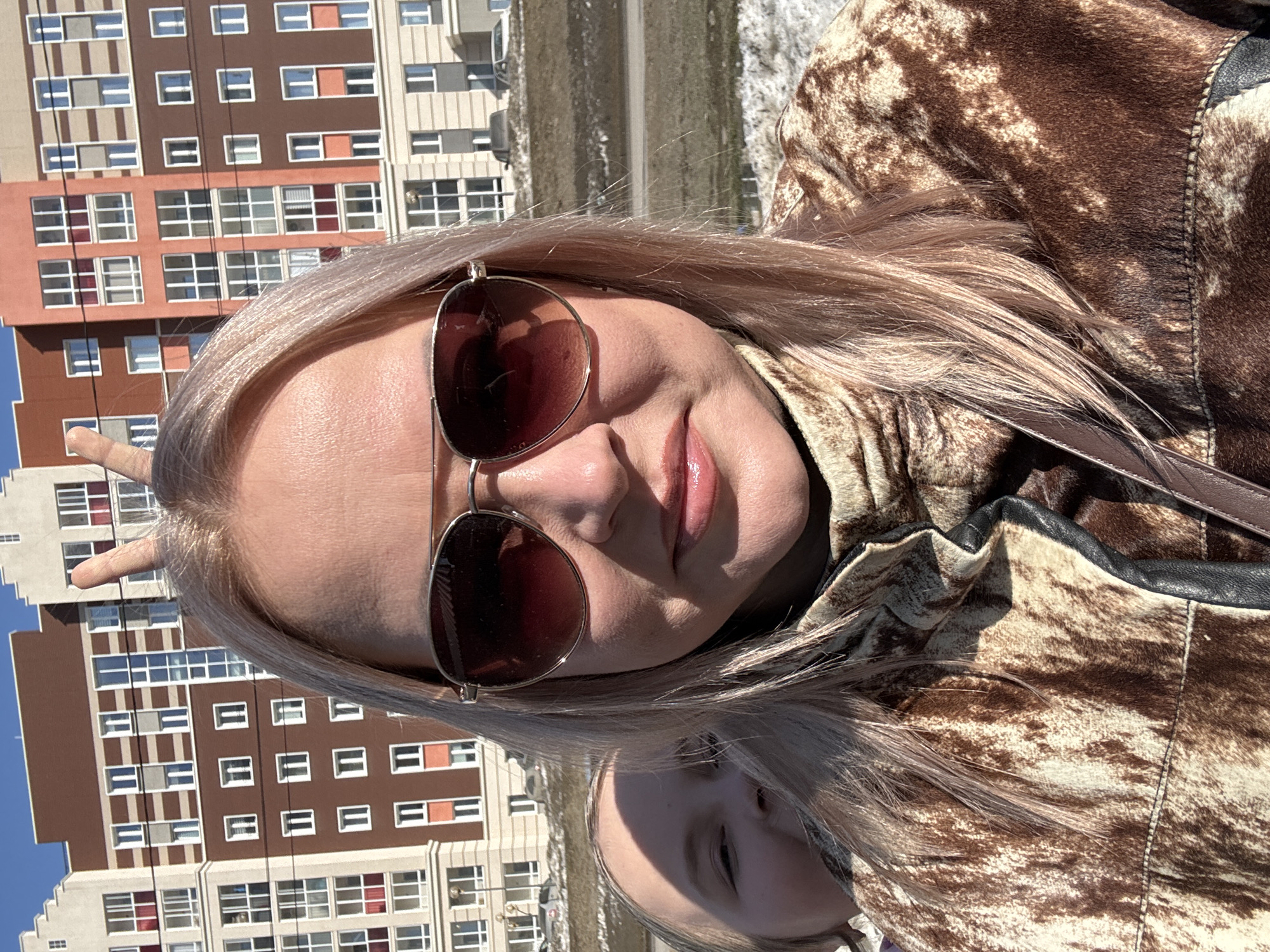 A woman is shown wearing sunglasses and a brown and beige patterned jacket. She is positioned in front of a tall, multi-story building in Pervomaysky, Russia. The building features rows of windows with a mix of beige, brown, and orange-red accents. The woman is making a hand gesture. Part of another person's face is visible in the bottom of the frame.