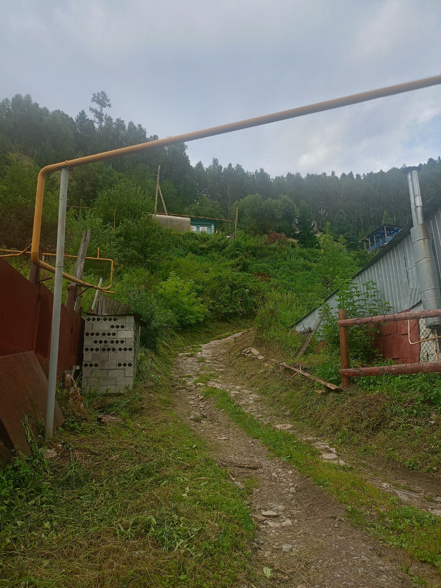 The image depicts a rural or semi-rural setting with a dirt road winding uphill through overgrown vegetation. A yellow pipe runs horizontally across the upper portion of the frame, supported by a metal pole on the left. To the left of the road, a rusty metal structure and a stack of grey cinder blocks are visible. The road itself is covered with stones and has tire tracks. On the right side of the road, a building with a corrugated metal roof and a large metal chimney is present, with a wooden fence in front of it. Further up the hill, dense green trees cover the landscape, and several small structures, including a green building, can be seen amidst the foliage. The sky is overcast. This scene is likely located in the city of Zlatoust, Russia, given the context provided by the user's request.