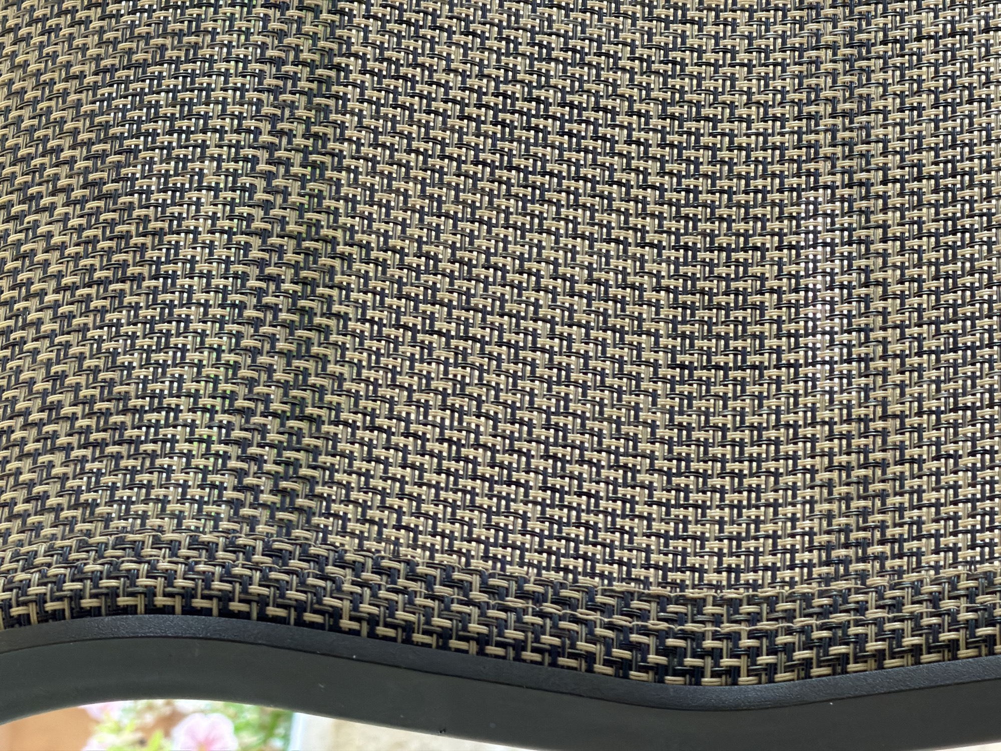 The image shows a close-up view of woven material on a chair. The material has a pattern of alternating light tan and dark blue threads, creating a textured surface. The weave appears to be a basketweave pattern. To the left of the woven material, a curved black edge of the chair is visible. In the background, out of focus, there are hints of greenery and possibly pink flowers, suggesting an outdoor setting. The image does not provide any information about the city of Kafr Yasif, Israel.