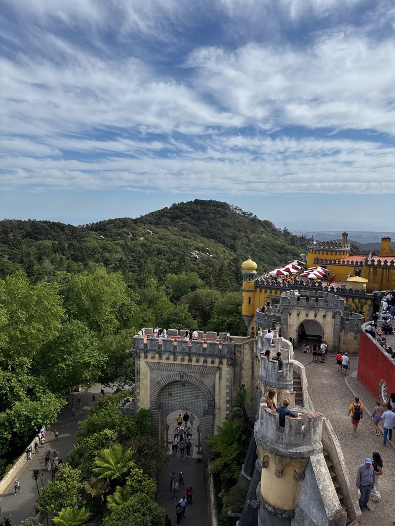 An eye-level, full outdoor shot depicts a partial view of the Pena Palace in Sintra, Portugal, with various people on the palace grounds. The scene shows portions of the palace architecture in the middle ground.
The left shows a stone archway with a metal gate and numerous people walking through it. The right side features a yellow tower and red wall. A staircase connects levels, and people are walking around and standing on the palace walls.
In the background, there's a densely forested mountain and a sky filled with wispy, horizontal clouds. The scene appears to be captured on a bright, sunny day.