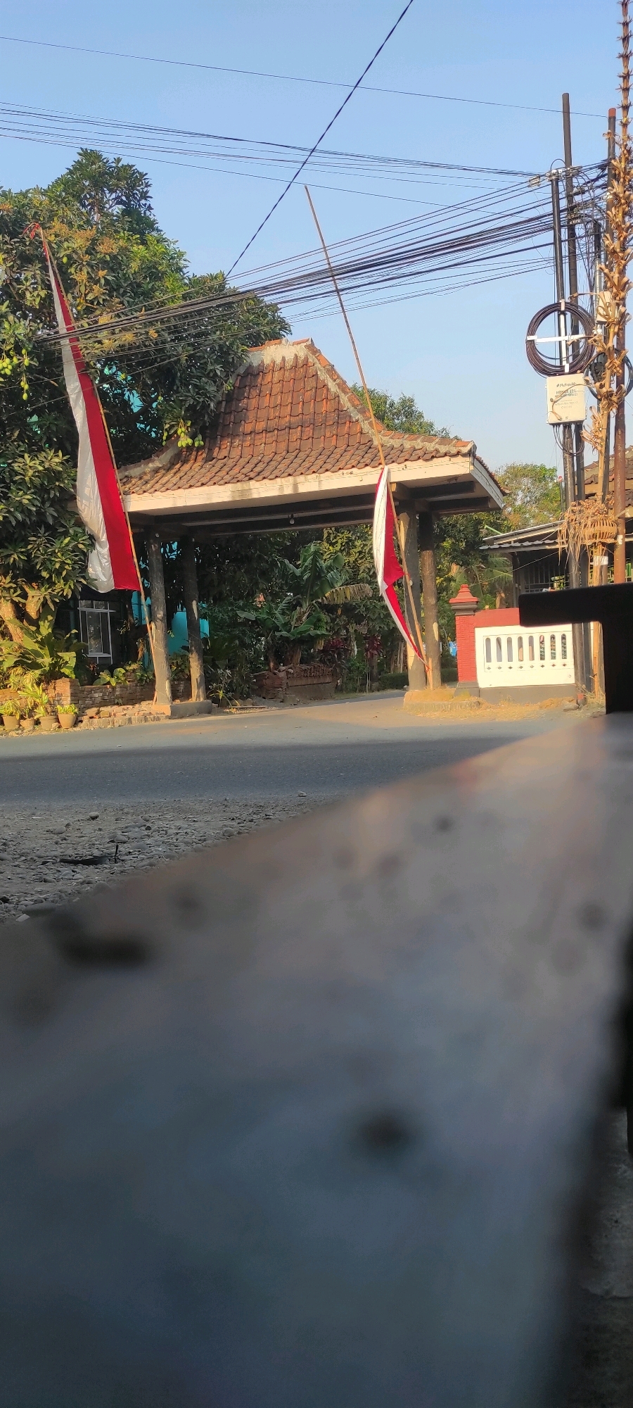 The image captures a street scene in Purwodadi, Indonesia, featuring a prominent gateway adorned with Indonesian flags. The gateway has a tiled, pitched roof supported by pillars, and is flanked by lush greenery and trees. Two red and white Indonesian flags are visible, one on the left and another on the right side of the gateway, fluttering in the breeze. A road runs through the gateway, with a paved area on the left and a darker asphalt road in the center and right. Electrical wires crisscross the sky above. To the right of the gateway, there's a white decorative wall with vertical slatted openings. The foreground of the image is dominated by a blurred, dark wooden surface, suggesting the photo was taken from a seated position behind an object. The sky is clear and blue, indicating daylight.