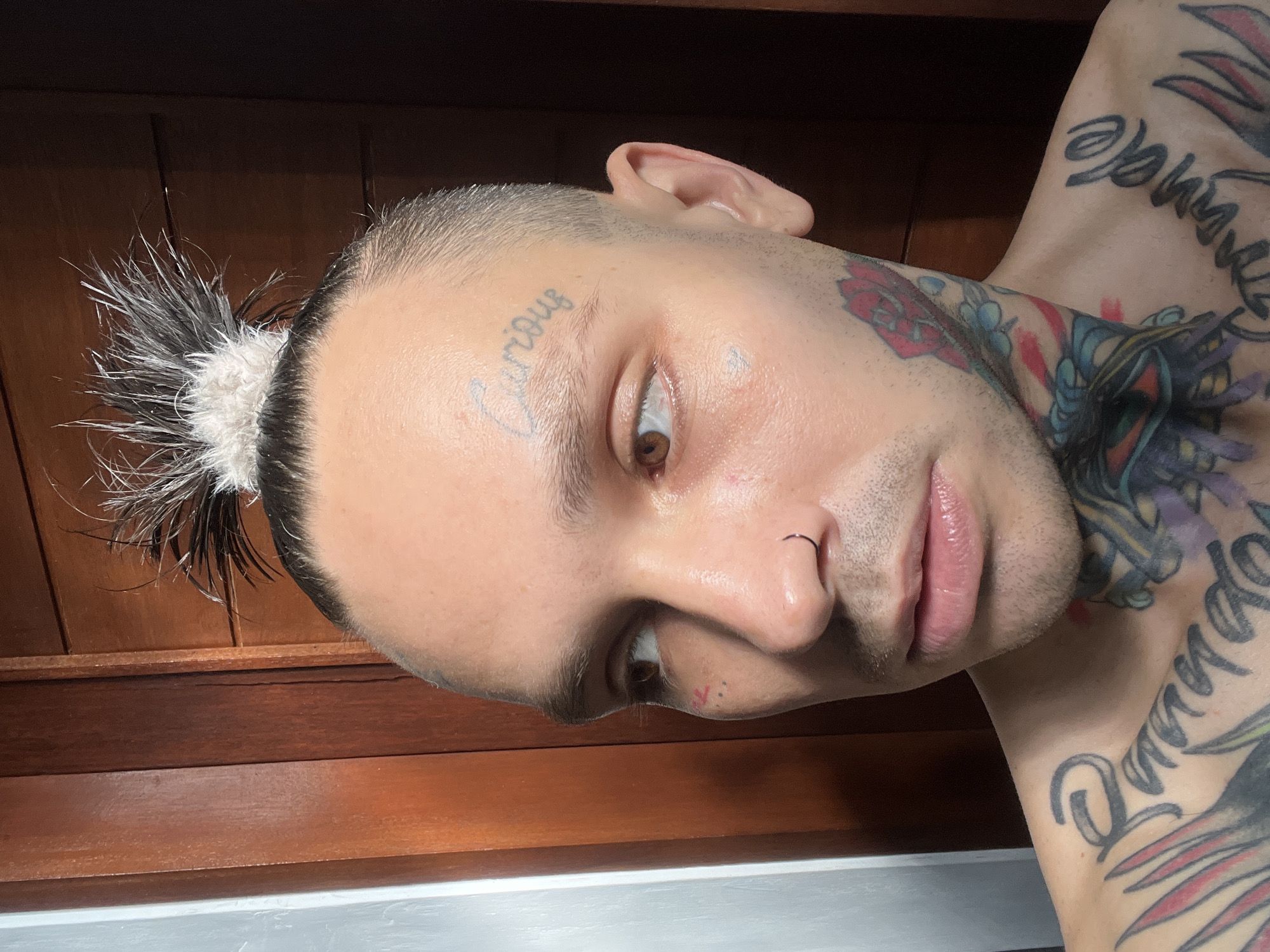 The image is a close-up, rotated portrait of a person. The subject has short, buzzed hair with a small topknot of white and black hair. They have a tattoo of the word "Curious" on their forehead. The person also has a nose ring and visible tattoos on their neck and chest, including what appears to be floral designs and script. The background consists of dark wood paneling. The lighting suggests an indoor setting, possibly with natural light. The subject's expression is neutral, with their eyes open and looking towards the camera. There are no clear indicators of time of day, weather, or specific location, beyond the indoor setting.