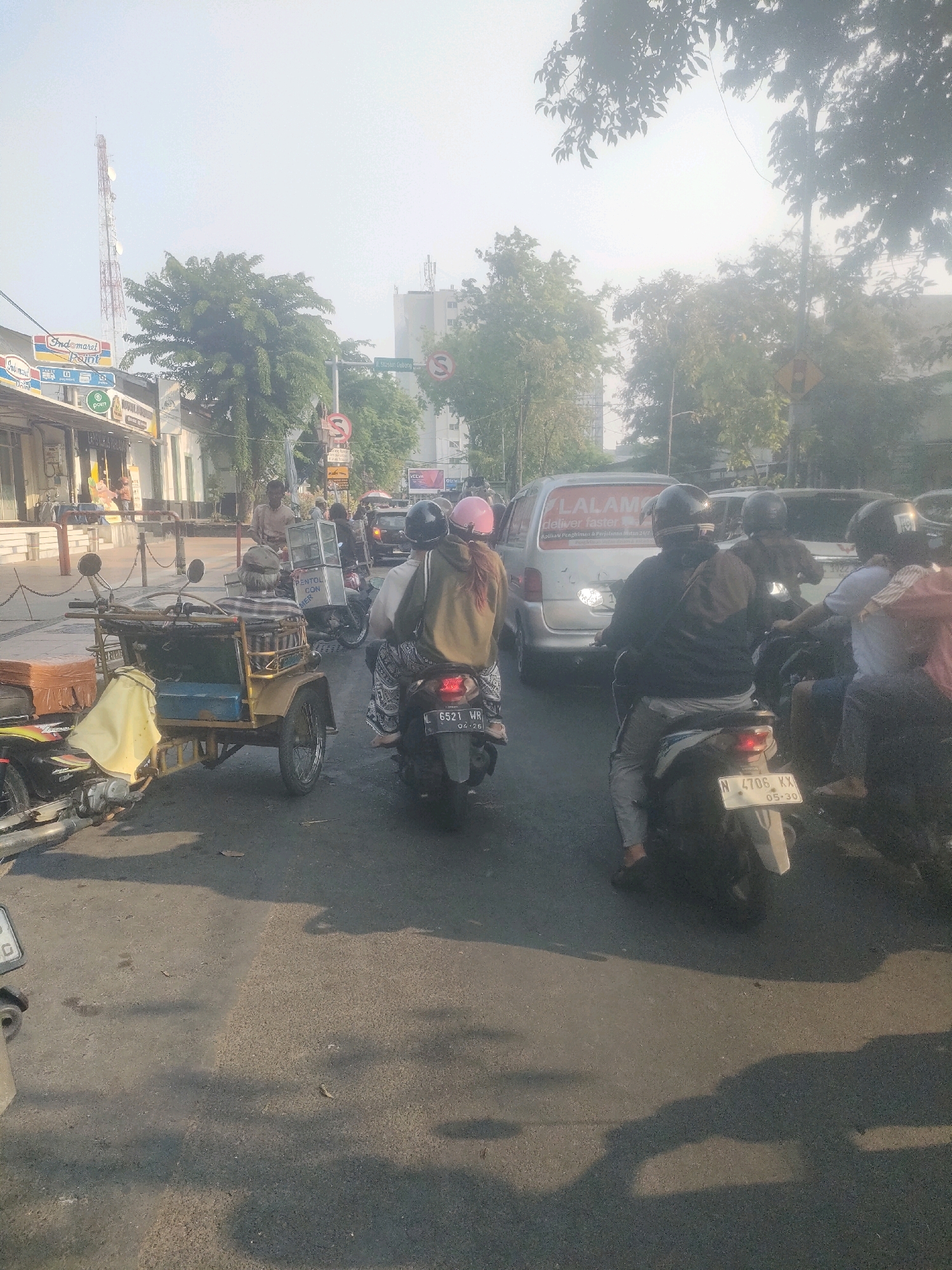 The image depicts a street scene in Surabaya, Indonesia, characterized by moderate traffic and a hazy, bright sky. In the foreground, a three-wheeled motorcycle, commonly known as a "becak motor," is parked, carrying a blue seat and a brown suitcase. Behind it, several motorcycles are in motion. One motorcycle has two passengers, a driver wearing a black helmet and a passenger with a pink helmet. Another motorcycle further ahead has a single rider in a black helmet and a grey jacket. A white van with text that reads "LALAMU" is visible in the middle ground, alongside other vehicles. The background shows buildings, trees, and a telecommunications tower, suggesting an urban environment. Street signs, including a "no U-turn" symbol, are present, indicating a regulated traffic area. The overall lighting suggests it is daytime, possibly late morning or early afternoon, with the sun casting shadows on the asphalt road. The air appears to have a slight haze, common in urban areas.