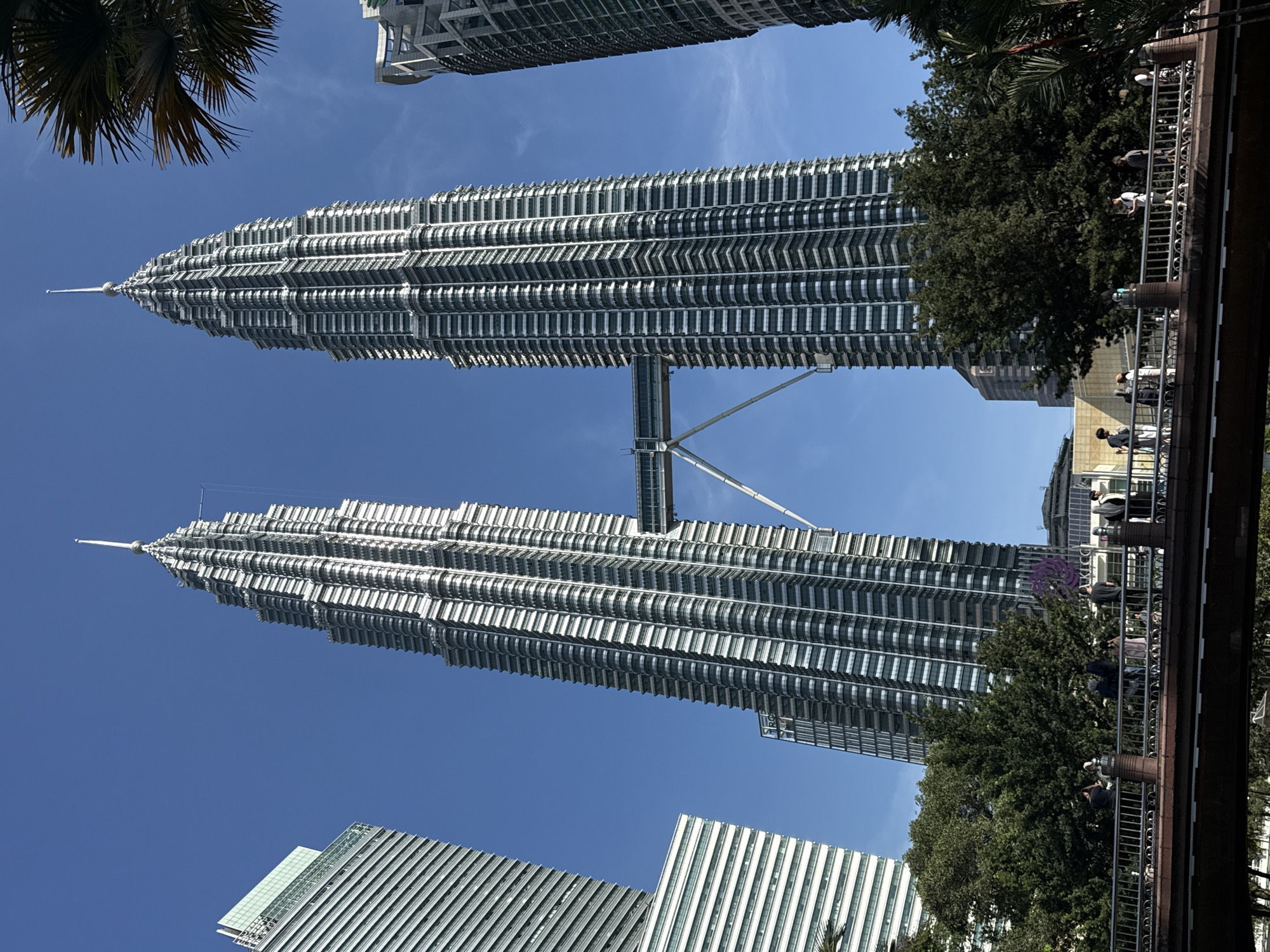 The image depicts the Petronas Twin Towers in Kuala Lumpur, Malaysia, viewed from a low angle. The two towers are conjoined by a skybridge. The sky is clear and blue. Palm trees are visible in the periphery. A pedestrian bridge is located on the right side of the image, populated with people. Other skyscrapers are visible in the background.