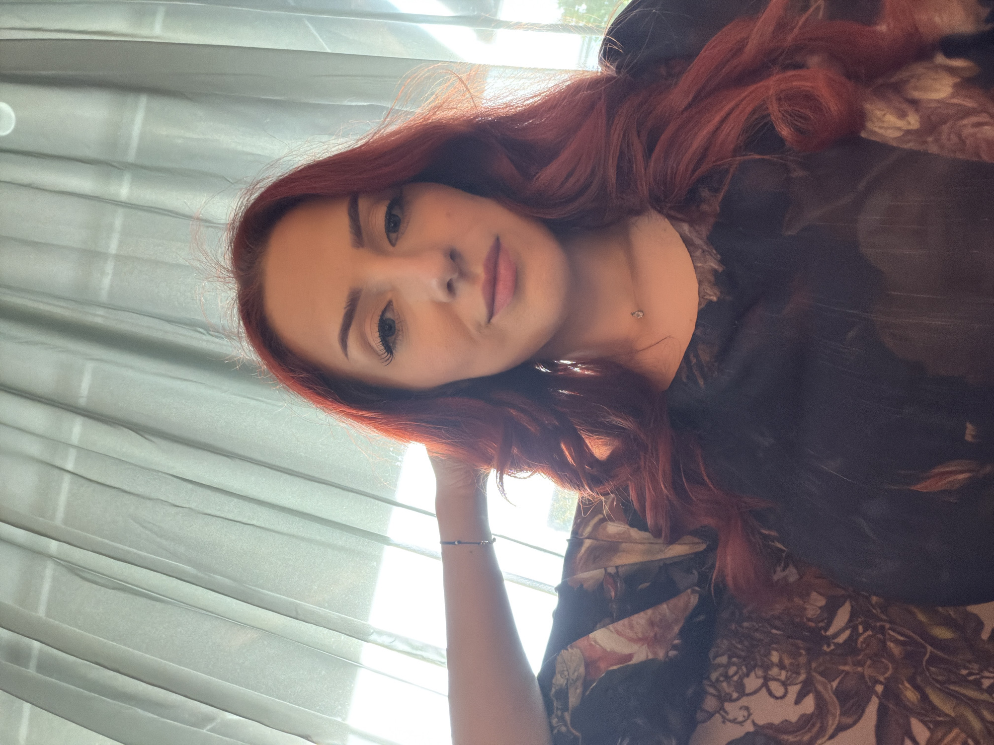 A medium shot of a woman with red hair. She is wearing a dark colored shirt with a floral pattern. She has a small necklace on, and a bracelet on her left arm. The background shows a window with light colored curtains. The photograph was taken inside. It is not possible to determine the location of the image as Sofia, Bulgaria, based solely on visual information.