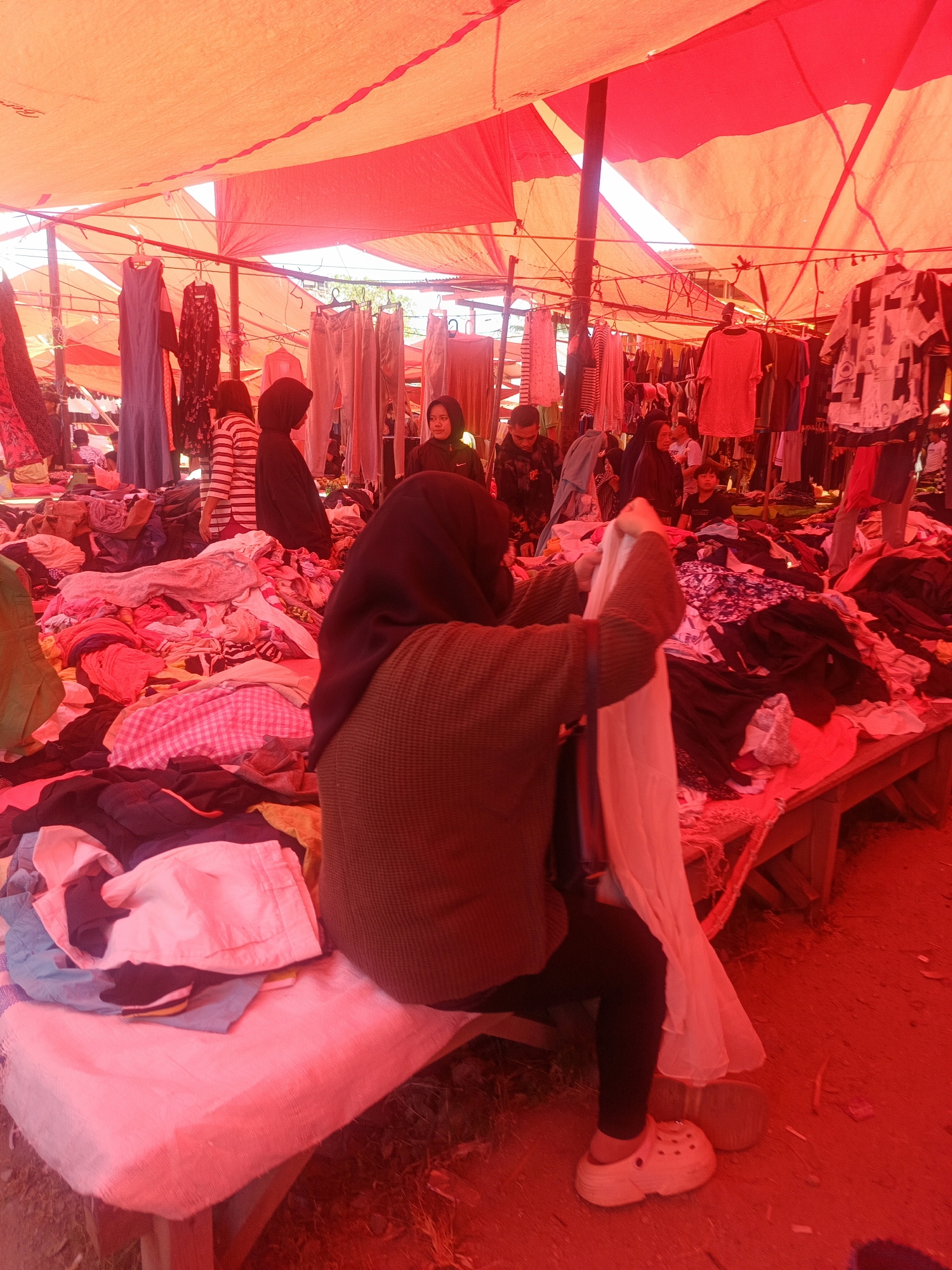 This image depicts an outdoor market stall in Gorontalo, Indonesia, likely a clothing market. The scene is dominated by a vibrant red tarp overhead, casting a strong red hue on everything. Several people are visible, some tending to the merchandise, while others appear to be browsing. Piles of clothing, including dresses, shirts, and pants, are displayed on tables and racks. The overall impression is one of a busy marketplace with an abundance of affordable clothing.