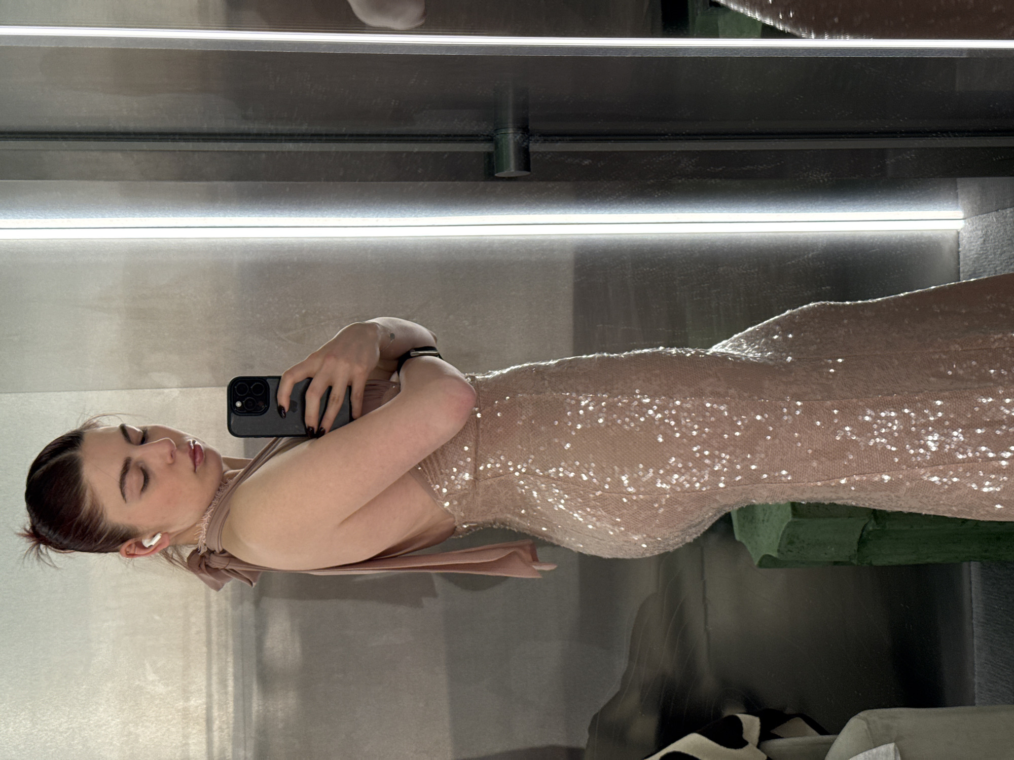 A person is seen in a mirror selfie in what appears to be a dressing room or fitting area. The individual has fair skin, dark hair pulled back, and is wearing a sequined dress with a bow detail at the back. They are holding a black smartphone in their left hand, presumably to take the photograph. The background is composed of mirrored surfaces and light panels, with glimpses of other objects in the reflection, suggesting a retail environment. There is no indication of the location being in Moscow, Russia, or any specific geographical location within the image.