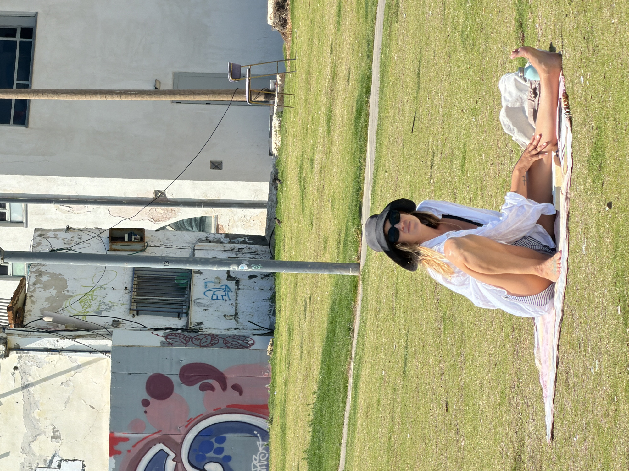 A woman wearing a white shirt and a black hat is sitting cross-legged on a blanket in a grassy area. Buildings with graffiti on the walls are visible in the background. The location appears to be Tel Aviv, Israel.