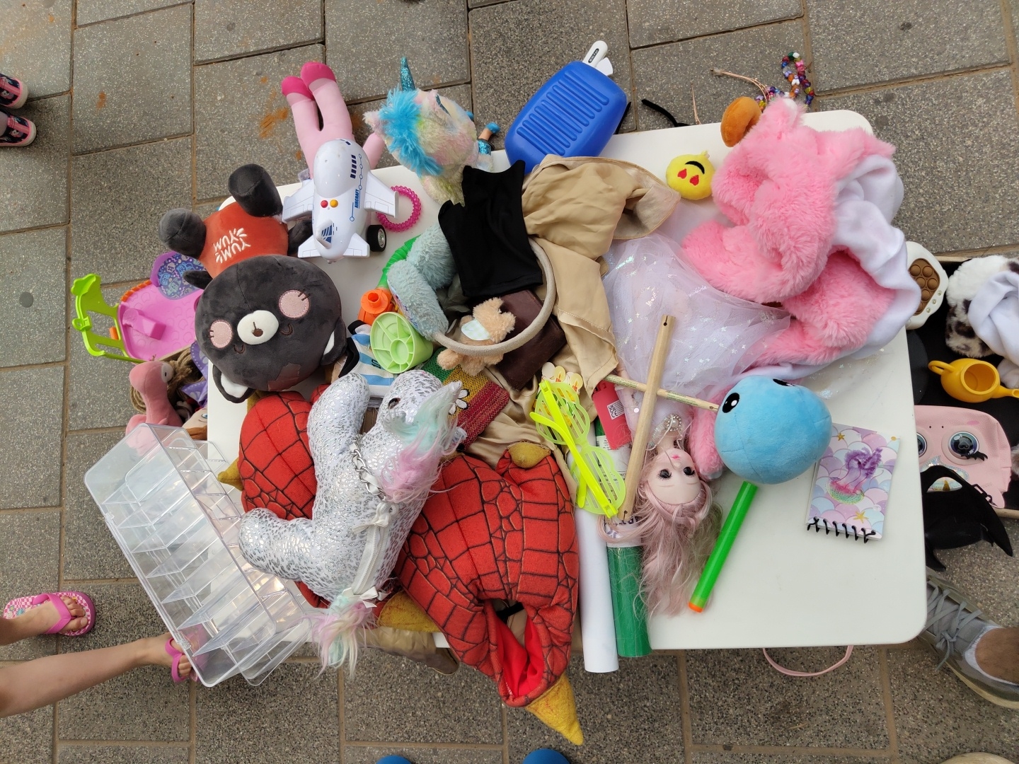 A top-down view reveals a cluttered white folding table on an outdoor stone-tiled surface, likely a sidewalk or patio. The table is covered in a variety of toys and other objects, including stuffed animals (a pink shark, a unicorn, a grey bear, and a red Spider-Man), a white toy airplane, a blue water bottle, and various other plastic toys. A doll with long pink hair lies near the center of the table. Also present are a small notebook, some pieces of cloth (tan, black, white tulle, and pink fleece), and a clear plastic storage container, which is partially open. At the edges of the frame are portions of people's legs and feet. To the left, a person wearing pink flip-flops stands near the table. At the top left, portions of two feet clad in children's shoes are visible. At the bottom right, a person wearing grey sneakers stands by the table. The location, inferred from the presence of a Hebrew-language newspaper on the table, is Tel-Aviv, Israel.