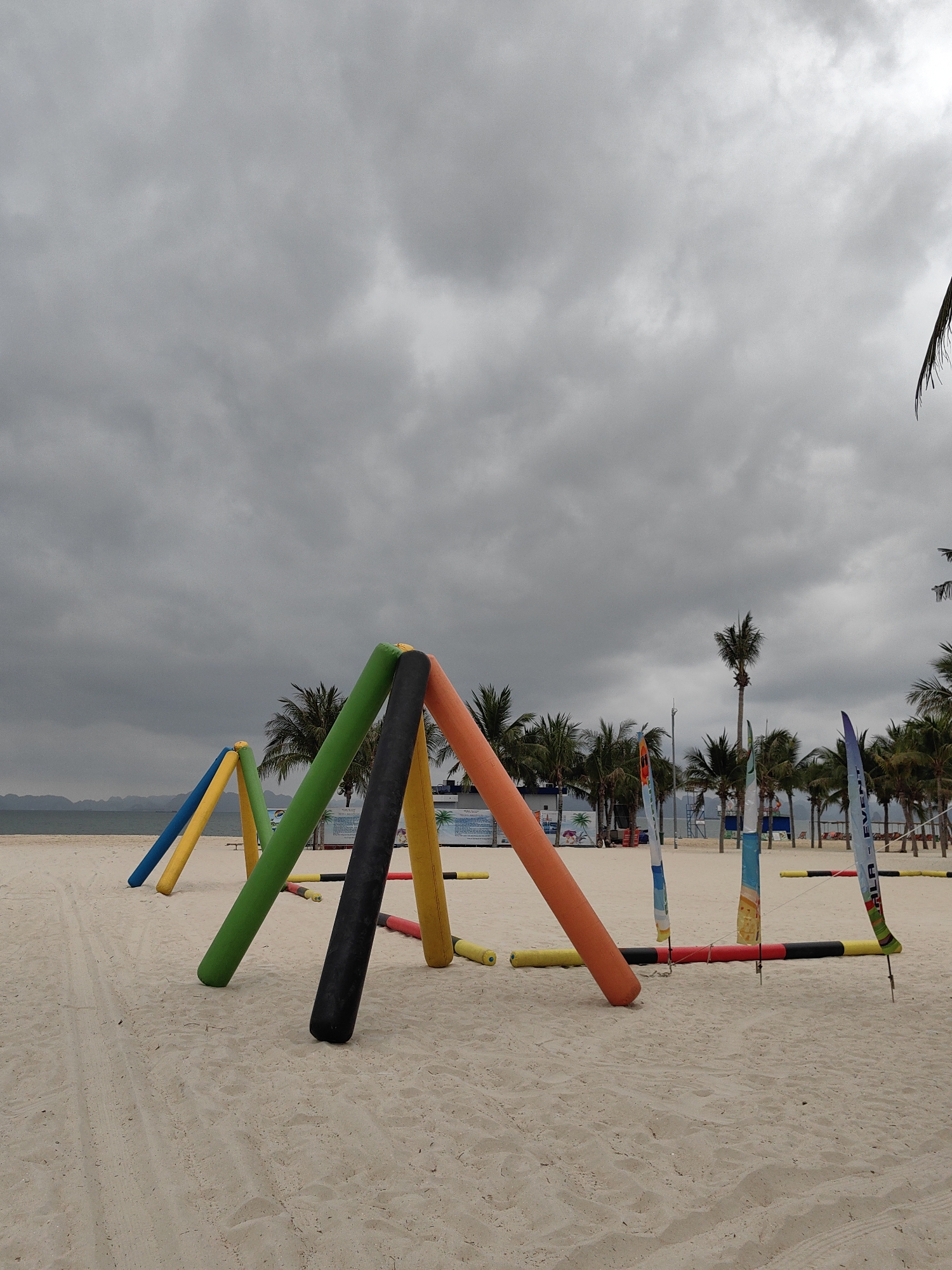 The image shows a beach in Bai Chay Ward, Vietnam, under an overcast sky. In the foreground, there are colorful inflatable arches and poles arranged on the sand. Behind them are several palm trees. Further in the background, there is a view of the ocean and indistinct landmasses. The sand shows signs of being traversed.