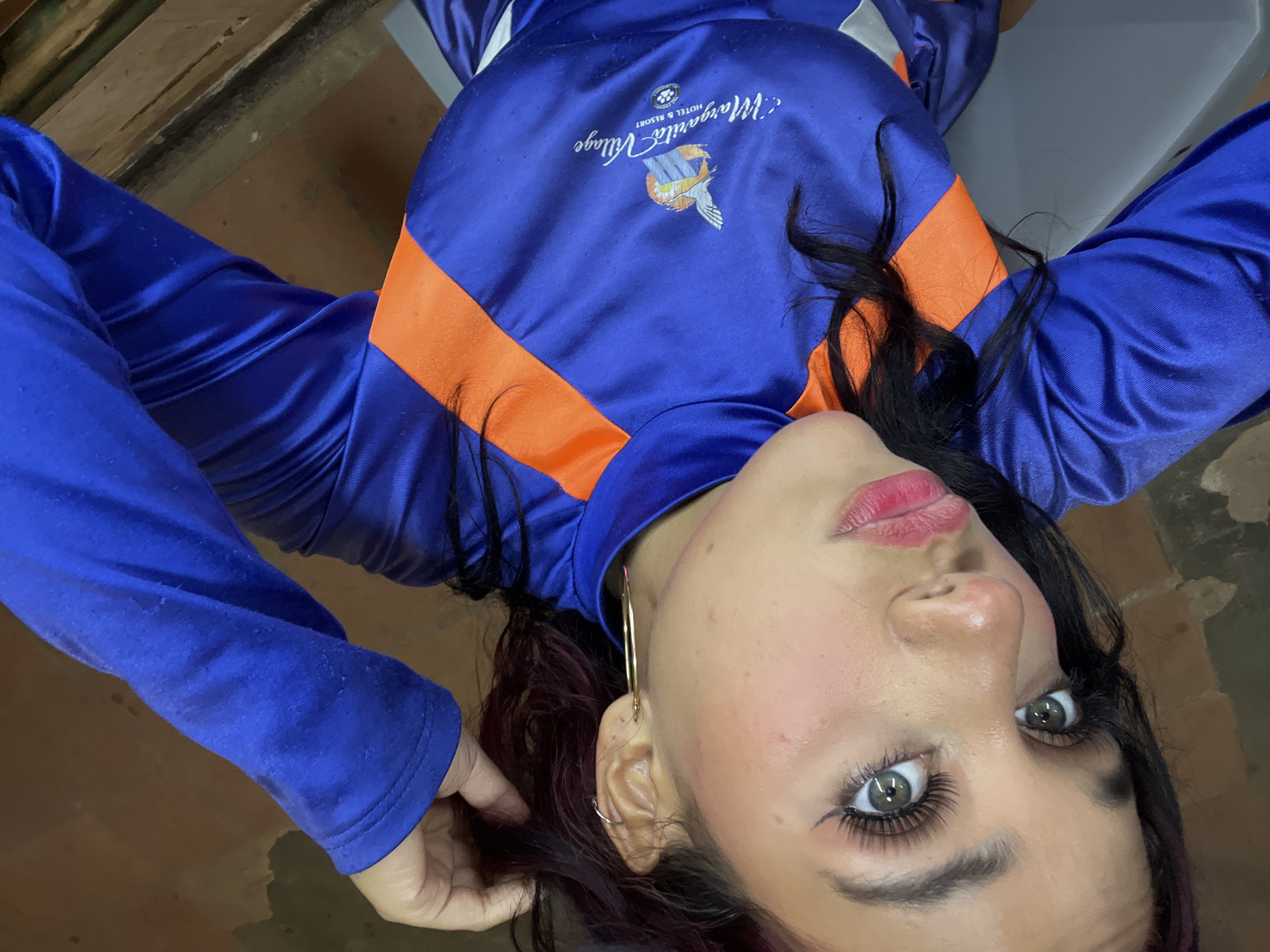 A selfie is taken by a young woman with dark hair and makeup. She is wearing a blue and orange long-sleeved shirt. The shirt has a logo on the front that reads "Margarita Village Hotel & Resort". The woman is looking at the camera with her left hand touching her hair. The background appears to be indoors with brick walls and wooden surfaces.
