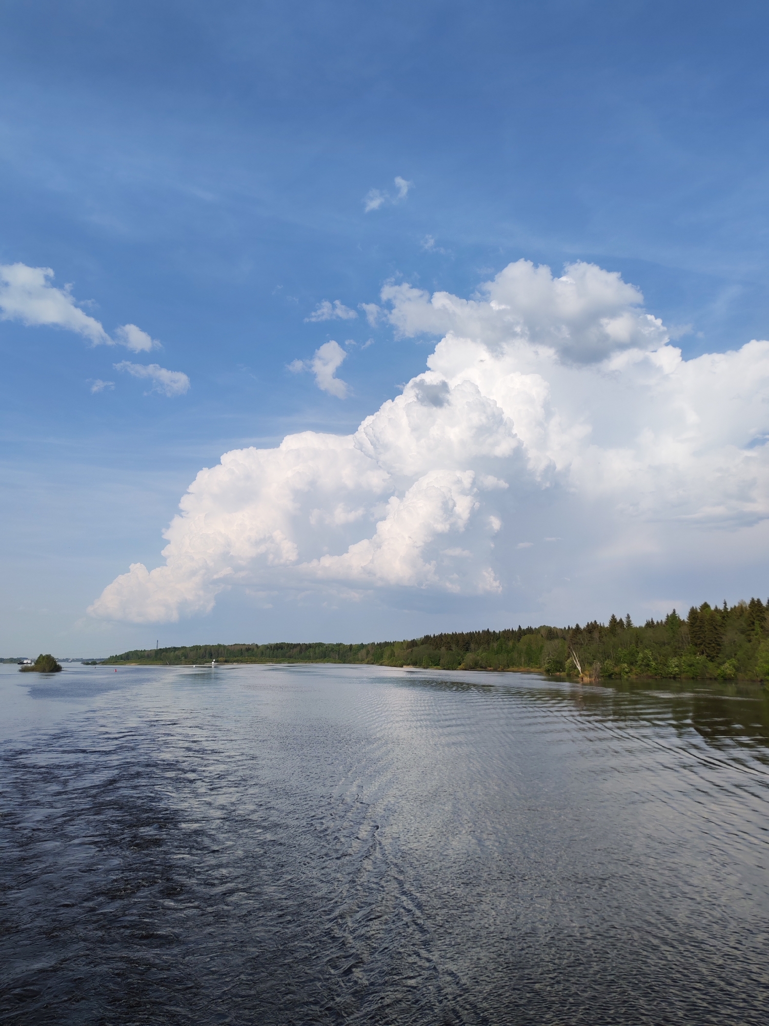 The image shows a body of water, likely a river or lake, with land visible on the right side and distant land on the left. The water's surface is mostly calm with some ripples. Dense green trees cover the land on the right. The sky is blue with large, puffy white clouds dominating the upper portion of the frame.
While the image may resemble a typical river view in a wooded area, without verifiable metadata or further information, it's impossible to definitively determine whether this photograph was taken in Sud'bitsy, Russia.