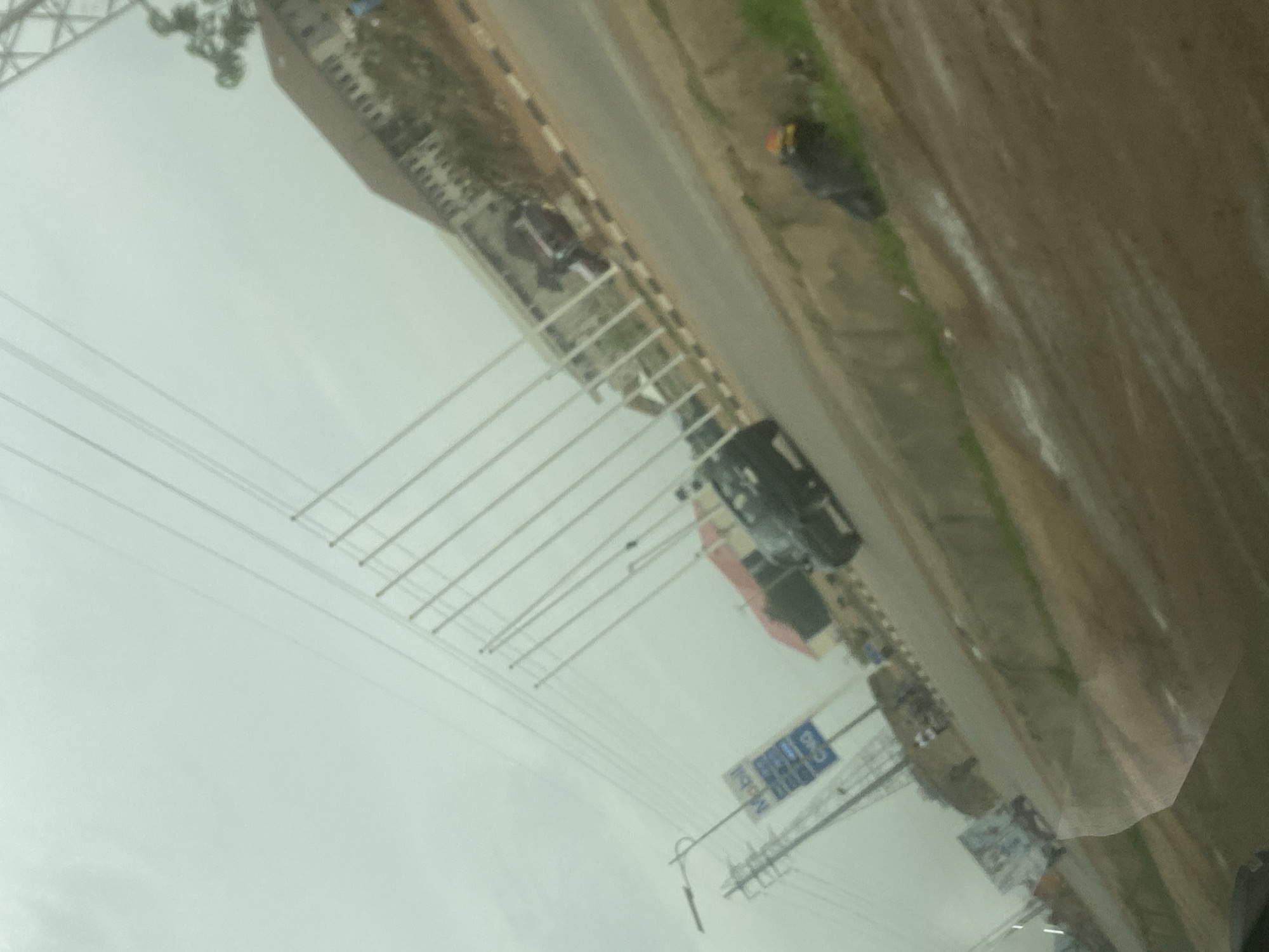 The image depicts a street scene in Abuja, Nigeria, taken from inside a vehicle, likely a car, given the visible portion of the dashboard. The road has asphalt pavement, with a building to the left and the land to the right. Vehicles including cars are on the road. Electrical poles are seen on the street with many electrical wires spanning above the road. There are signposts on the street side. The sky is overcast, casting a dull light over the scene. The road appears slightly wet, suggesting recent or ongoing rain. The angle and composition suggest the photo was taken quickly, and the focus is on the general environment rather than any specific subject.