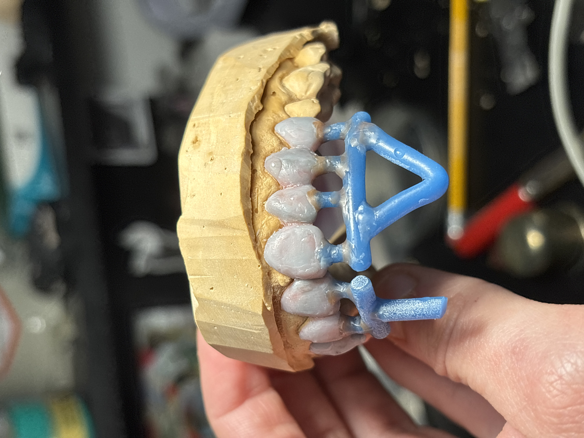 A human hand, visible in the lower-left, firmly grasps a light tan dental arch model, likely crafted from gypsum or plaster. The model depicts a maxillary arch. Attached to the anterior portion of this model are eight dental wax-ups, appearing light grey with subtle pinkish hues near the presumed gingival areas, resembling prepared prosthetic teeth. These wax-ups are supported by a textured, light blue resin framework, consistent with a sprue system used for dental casting or pressing. The background is out of focus, revealing a dark workbench or surface. Identifiable blurred objects include a yellow pencil, a red tool handle, and various containers, one with a teal and white label, indicative of a dental laboratory setting in Sofia, Bulgaria.