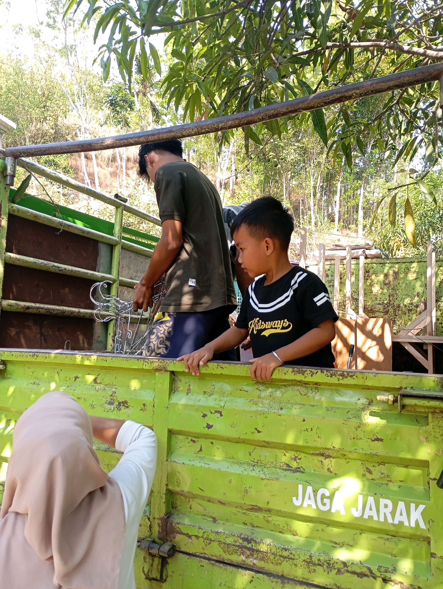 In Ketepung, Indonesia, a truck bed, painted lime green and showing signs of wear and rust, is the central focus of the image. Two young boys are positioned in the truck bed. The boy on the right, wearing a black t-shirt with white stripes and yellow lettering that reads "Kidsways," leans on the edge of the truck bed. To his left, an older boy, wearing a dark olive green t-shirt and blue shorts with a yellow pattern, is in the process of handing a metal object, possibly a decorative fence component, out of the truck. In the foreground, partially visible from behind, a person wearing a beige hijab and a white long-sleeved shirt reaches into the truck bed. The background reveals a lush, sun-drenched natural environment with trees and foliage. The text "JAGA JARAK" (meaning "keep distance" in Indonesian) is stenciled in white on the side of the truck bed.
