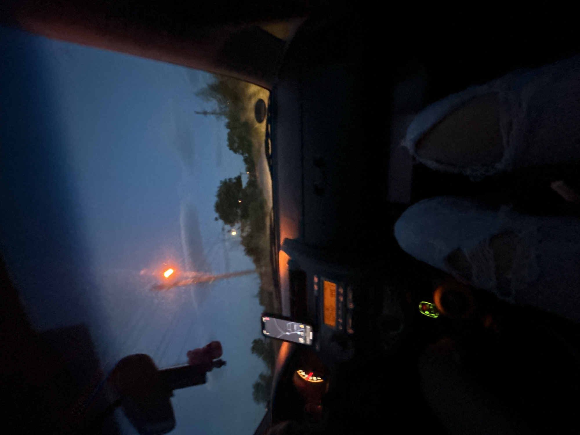 The image depicts the interior of a car driving at night. Outside the windshield, a road is visible under a dark blue sky with a single orange streetlight. In the foreground, the car's dashboard is partially visible, with a phone mounted on a holder, displaying a map. The driver's legs, clad in ripped jeans, are also visible. The image does not provide sufficient information to confirm the location is Ovchepoltsi, Bulgaria.