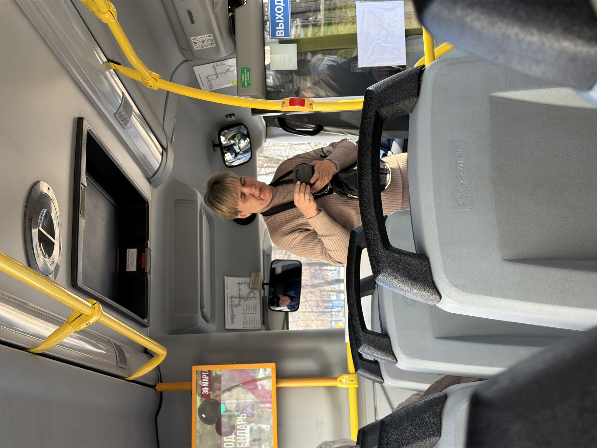 Here is a forensic description of the image:
The photograph depicts the interior of a public bus. A woman is visible in the middle of the frame. She is light-skinned with short blonde hair, wearing a light brown sweater, and has a black camera hanging around her neck. She is holding and looking at the camera, apparently adjusting or checking it.
The bus interior is mostly gray with yellow handrails. There are several signs with text in Russian. Some signs mention "30 MAPTA (March 30)" and "Выход (Exit)". The "СОТЕКС" brand is visible on the side of the back of the seat. The scene appears to be from the city of Chaikovsky, Russia, based on the signage. The photograph appears to have been taken from a low angle, possibly from a seated position in the bus.