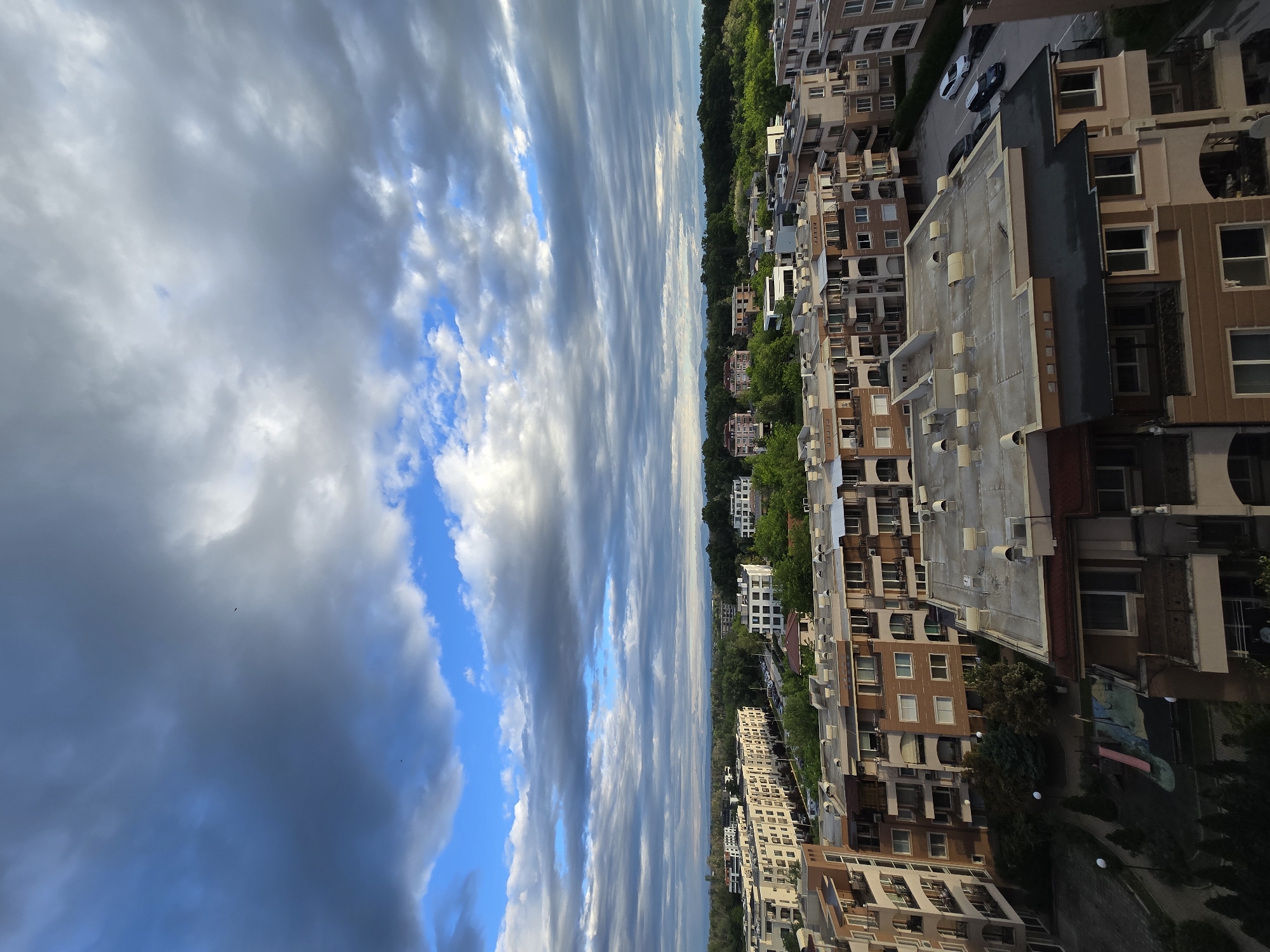 Here is a detailed description of the image:
The image shows a cityscape under a dramatic sky filled with streaky clouds and patches of blue sky. The perspective is likely from an elevated position, looking across a neighborhood with multiple buildings.
Buildings:
The majority of the lower half of the image is dominated by multi-story buildings, mostly constructed of brick or tan-colored materials. There is visible variety in the architectural style, with some buildings featuring balconies, varying window designs, and roof structures. Some roofs are flat and have visible equipment (possibly HVAC units).
Environment:
A line of trees separates some of the buildings. This suggests a green belt or park within the residential area.
The sky above takes up a significant portion of the image and contains a variety of cloud formations.
Overall:
The image conveys a sense of urban living but also indicates that the neighborhood is near green spaces. The weather seems unsettled.