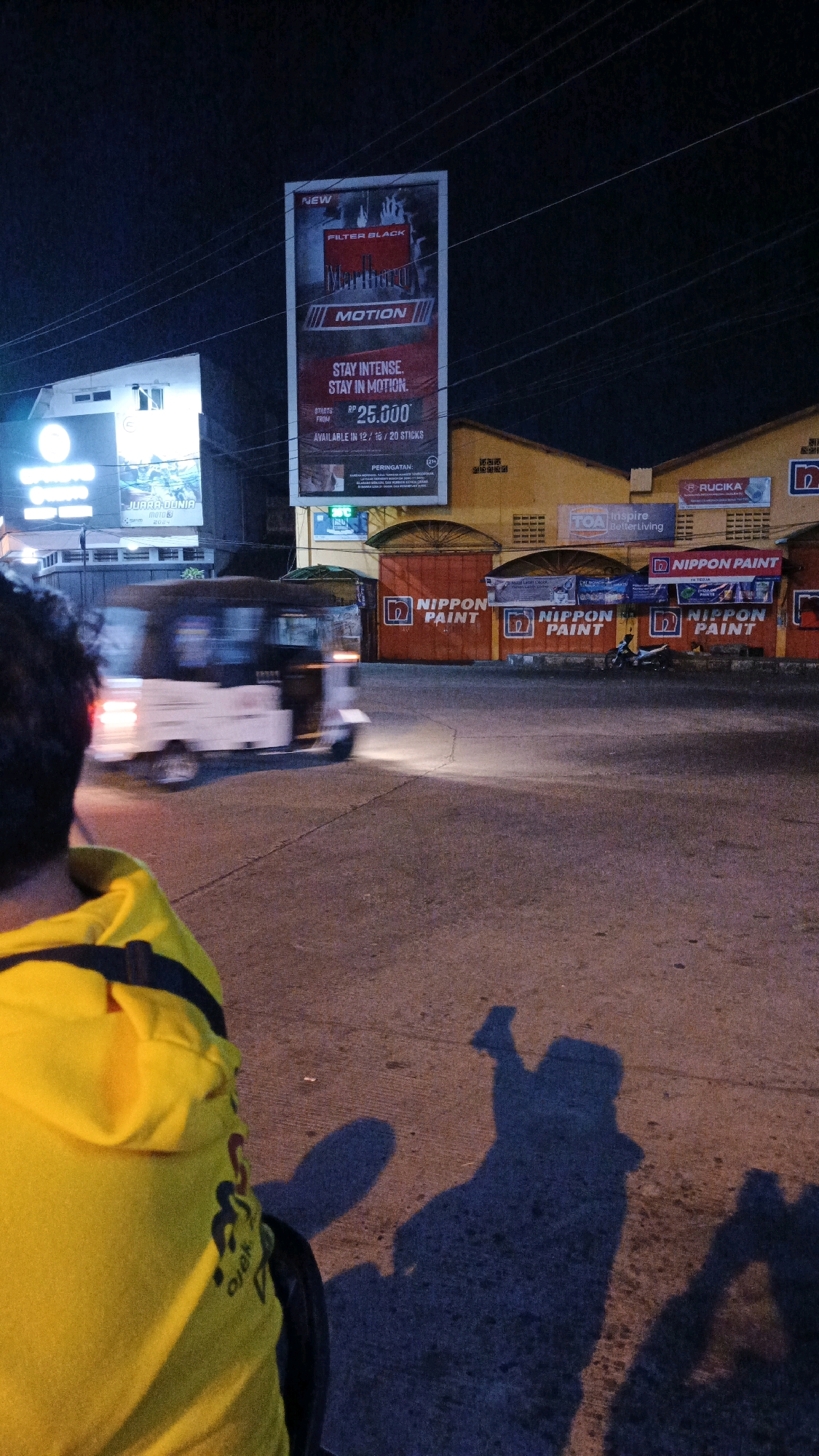 An outdoor, night-time scene captures a street in Surakarta, Indonesia. The foreground shows the back of a person wearing a bright yellow hooded jacket with black straps. Their shadow, along with others, is cast on the asphalt road. A three-wheeled vehicle, possibly an auto-rickshaw, is blurred by motion in the mid-ground, its headlights illuminated.
In the background, buildings line the street. A prominent billboard advertising "MARLBORO MOTION" hangs above a building with yellow walls and a reddish-orange corrugated metal facade. Several large orange garage doors with "NIPPON PAINT" branding are visible, along with smaller signs for "TOA Inspire Better Living" and "RUCKA". Another building on the left has a brightly lit white facade with a sign displaying "JUARA DUNIA" and the "Milo" logo. The overall impression is a commercial street at night with active traffic and illuminated signage.