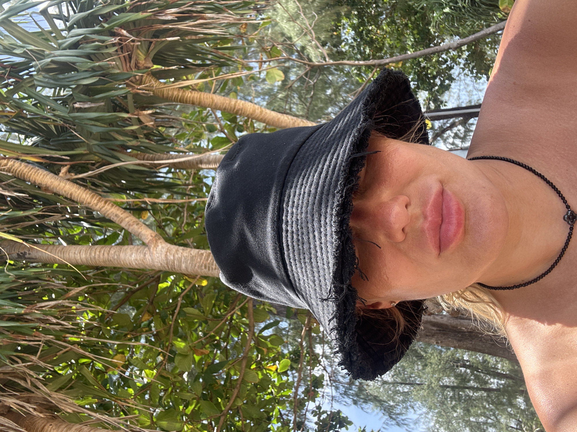 A close-up, over-the-shoulder shot captures a person wearing a black bucket hat with a frayed brim. Their face is partially obscured by the hat, with only their lips and a portion of their cheeks visible. They are wearing a dark beaded necklace with a floral pendant. The background features lush green foliage and palm trees, indicative of a tropical environment, likely in Choeng Thale, Thailand. The sunlight creates dappled shadows, suggesting an outdoor setting during the day. The person's skin appears tanned, hinting at a warm climate.