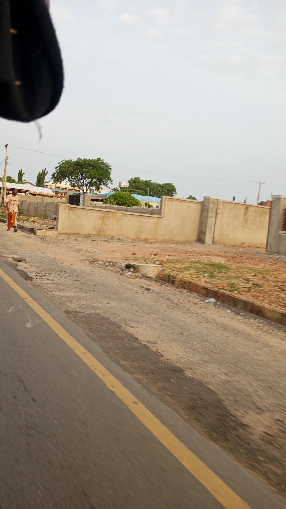 The image shows an urban scene in Lokoja, Nigeria. The foreground features an asphalt road with a yellow dividing line. The road is adjacent to a partially developed plot of land with exposed soil and sparse vegetation. A light-colored concrete wall borders the plot. A man wearing a patterned shirt and orange pants is walking along the road. Overhead power lines are visible in the background, along with trees and low-rise buildings. The sky is overcast.