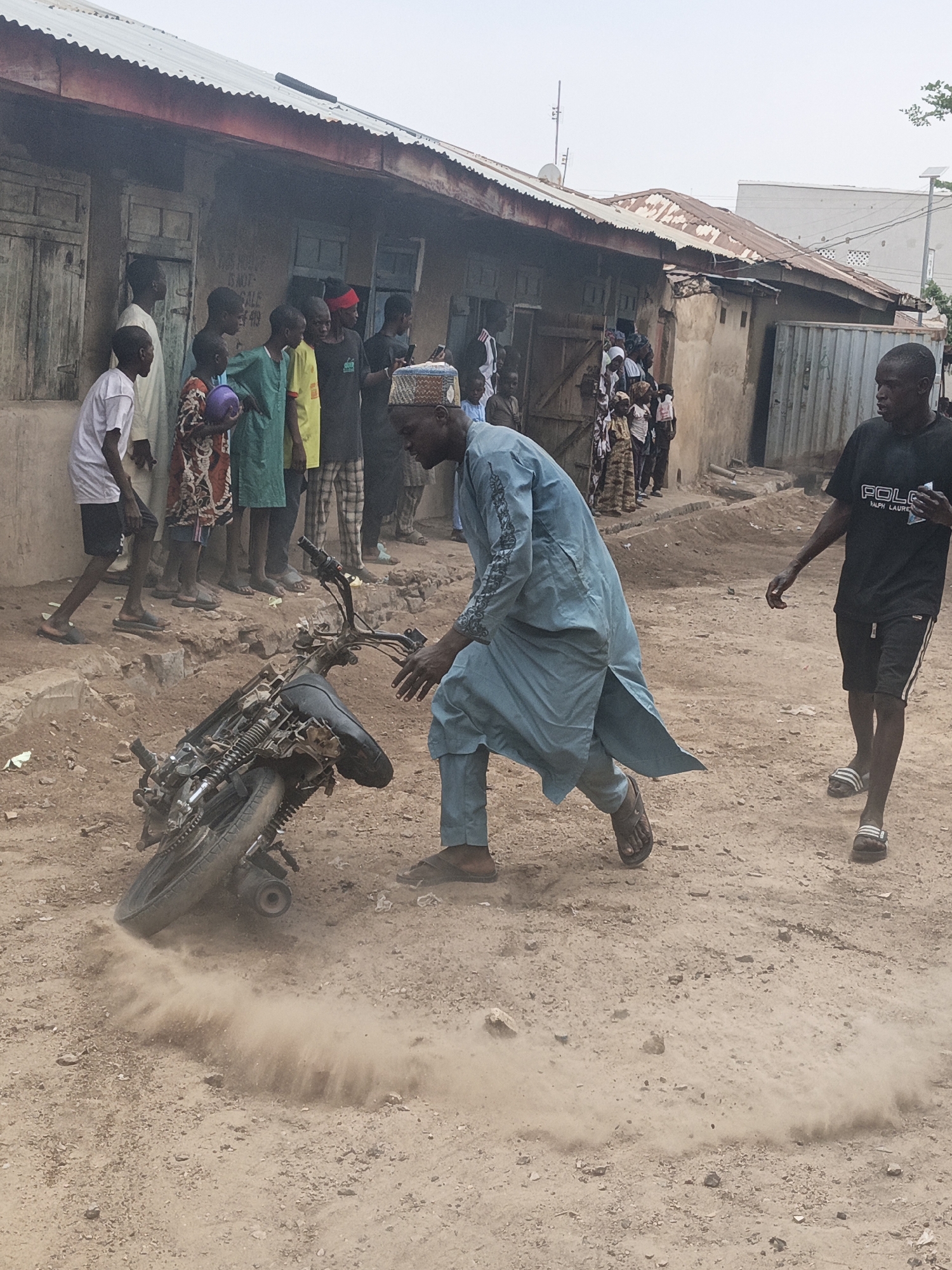 In Gwagwarwa, Nigeria, a dusty, unpaved street is the setting for a scene of commotion. A motorcycle lies on its side, kicking up a cloud of dust, suggesting it may have recently crashed or been hastily dropped. Two men are the primary focus. One man, dressed in a flowing light blue robe and dark pants, is stooped over the motorcycle, his hands extended as if to steady it or examine it. He wears dark sandals. To his right, another man in a black t-shirt with "POLO" and "RALPH LAUREN" printed on it, and dark shorts, walks away from the motorcycle, holding a small white object in his right hand. He also wears dark sandals. Behind them, a group of onlookers, mostly young males and children, are gathered, observing the situation. They stand in front of a row of weathered, attached buildings with corrugated metal roofs. The architecture suggests a humble, possibly older, part of the city. The overall atmosphere appears to be one of disruption and public interest.