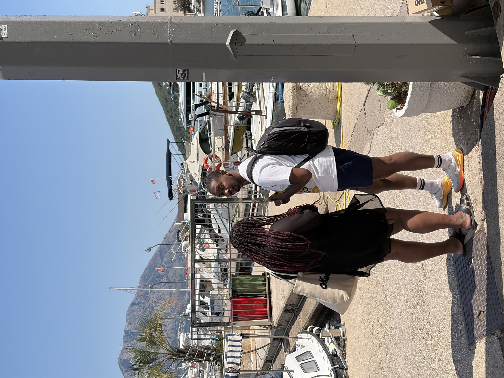 A man and a woman are standing on a paved walkway in a marina on a sunny day. The woman, with long, dark braided hair and wearing a black dress, stands facing away from the camera, holding a large tote bag in her left hand. The man, wearing a white t-shirt, navy shorts, white socks with black stripes, and bright yellow and white athletic shoes, stands facing the camera, looking slightly to his left. He has a black backpack on his right shoulder. Behind them, numerous boats of various sizes are docked, and in the background, mountains covered in greenery rise under a clear blue sky. A tall, gray utility pole is prominently positioned to the right of the frame, with stickers on its surface. The scene appears to be in Budva, Montenegro, as indicated by a sticker on the utility pole that reads "BUDVA MONTENEGRO".