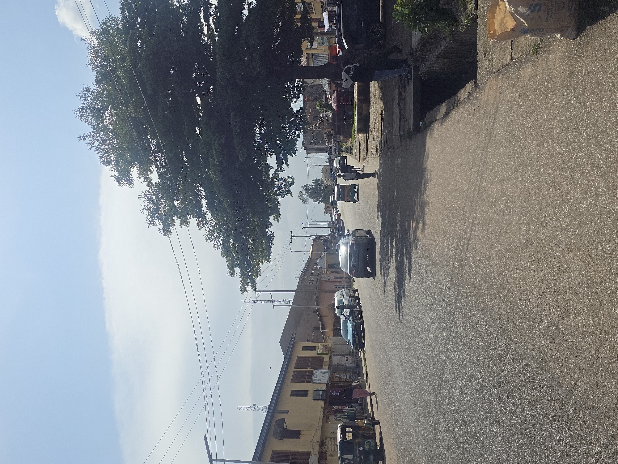 The image depicts a street scene in Jos, Nigeria, under a bright, partly cloudy sky. The perspective is from the side of the road, looking forward. On the left, a row of yellow buildings with balconies and shops line the street. A three-wheeled auto-rickshaw is parked on the left side of the road. Cars, including a dark sedan and a light-colored SUV, are driving on the asphalt road, with shadows cast by overhead structures and trees. On the right side of the road, a large tree with dense foliage dominates the foreground. People are visible on the sidewalk and crossing the street. Further down the road, more buildings and vehicles can be seen, along with utility poles and antennas. The environment appears to be a bustling urban area with commercial activity. A bag of cement is visible on the sidewalk in the lower right corner.
