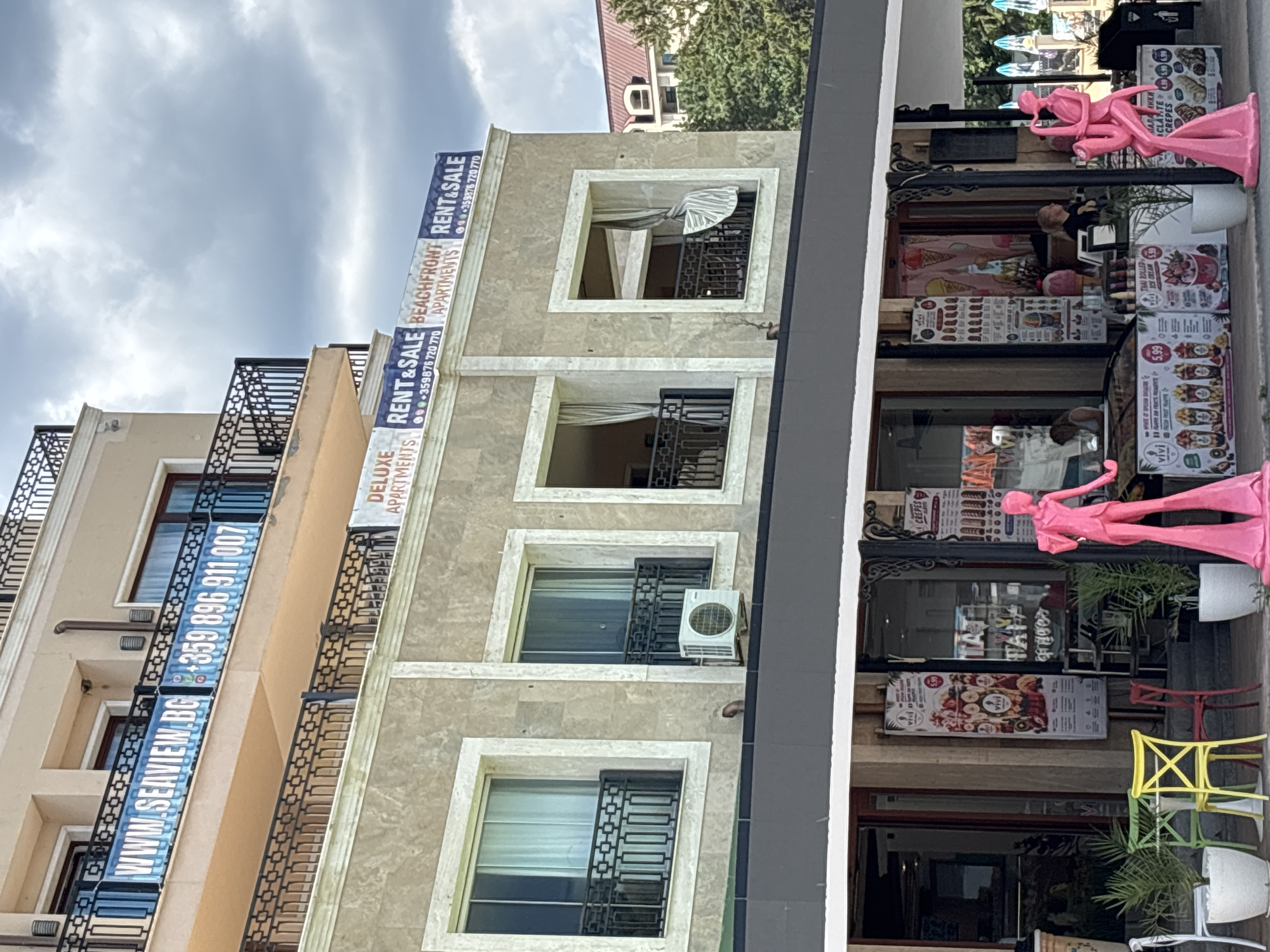 The image depicts a street-level view of a building in Varna, Bulgaria. On the upper floors, banners advertise "WWW.SEAVIEW.BG" and phone numbers, along with "DELUXE APARTMENTS RENT & SALE" and "BEACHFRONT APARTMENTS RENT & SALE." The building has balconies with black railings and cream-colored stone facades.
Below the upper floors, the scene transitions to a row of shops. A prominent feature is a row of colorful displays advertising food items, including "VIVI" branded crepes and ice cream, with prices visible. Pink mannequins, one in a tuxedo and the other playing a saxophone, are placed outside the shops as decorative elements. There are outdoor tables and chairs in front of the establishments, some with potted plants. People are visible inside the shops, seemingly serving customers. The sky above is cloudy. The overall impression is a commercial area, likely a tourist destination, given the advertising for apartments and beachfront properties.