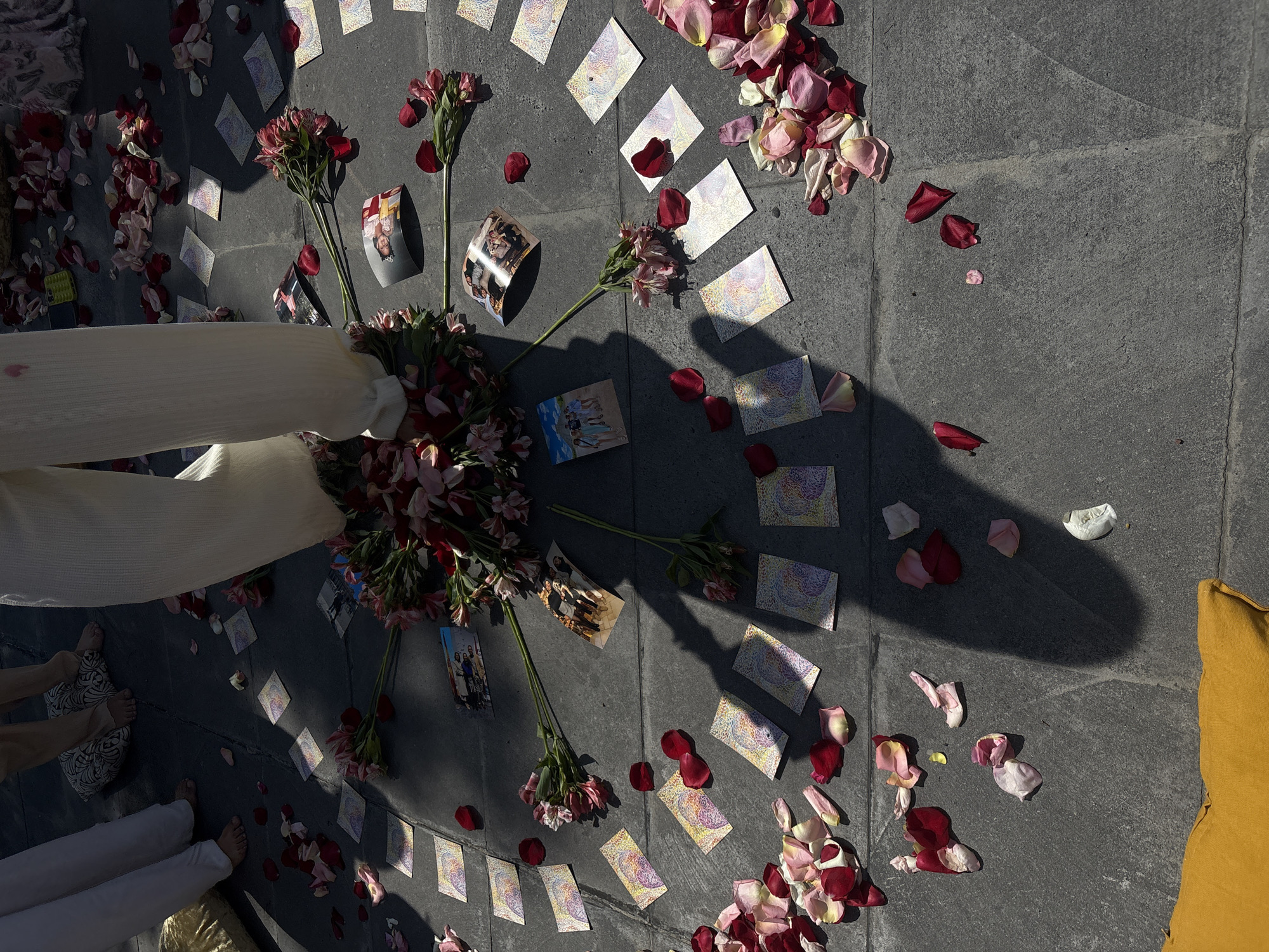 A top-down view depicts a ritualistic or memorial arrangement on a paved surface, likely outdoors in Puebla, Mexico. A semicircle is formed by scattered playing cards, red and pink rose petals, and several floral bouquets interspersed with printed photographs. A pair of legs dressed in white pants and a sheer garment, positioned on the left side of the image, suggests a human presence and possible involvement in the setting up of the arrangement. The arrangement sits on dark, textured paving stones. A shadow is cast on the right side of the image, close to a yellow fabric piece.