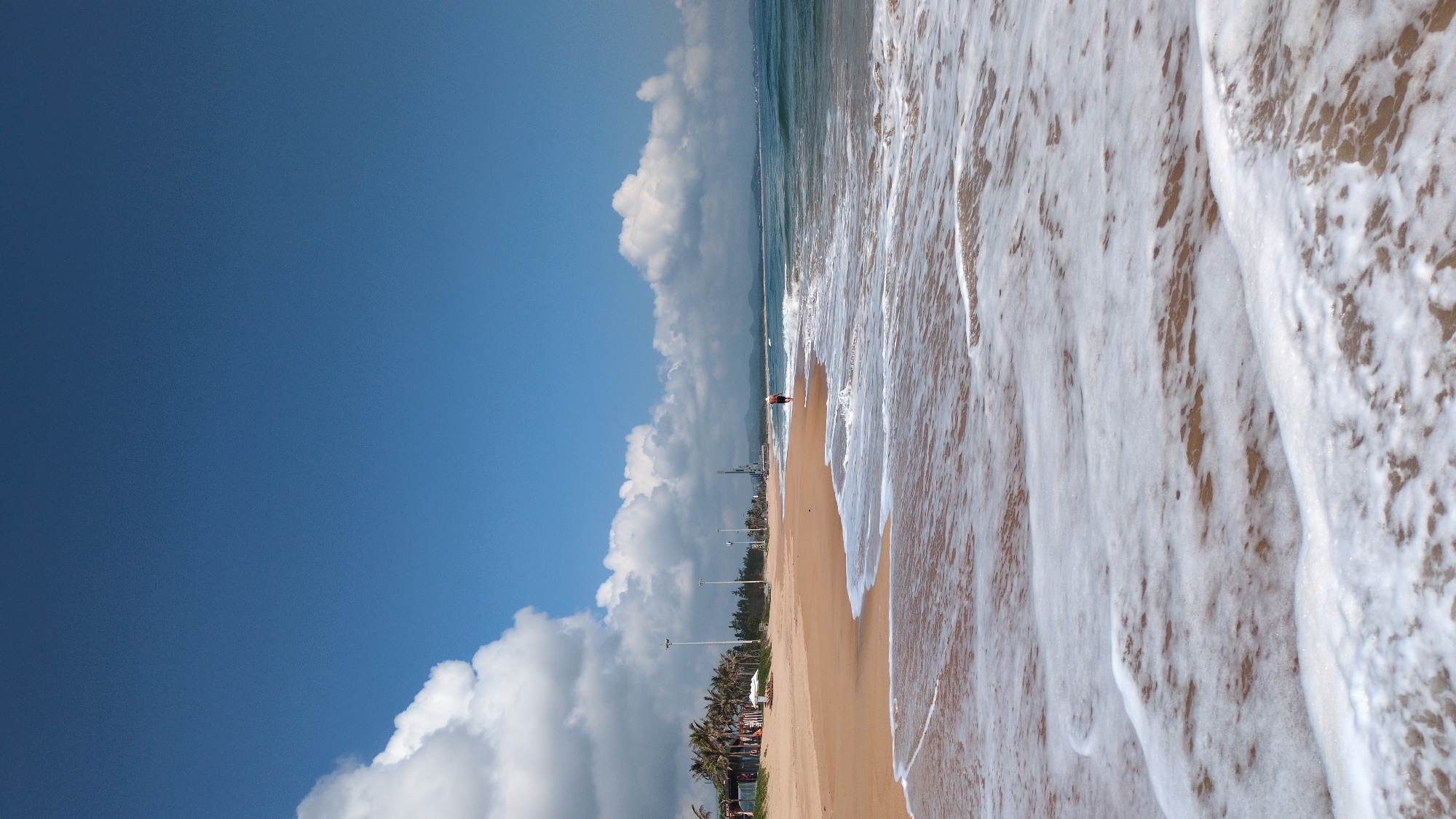 This image displays a daytime coastal scene on a sandy beach in Thành phố Tuy Hòa, Vietnam. In the immediate foreground, white seafoam from receding waves covers wet, light brown sand. The midground features a wide expanse of dry, light brown sand leading towards a line of palm trees and other green foliage. One individual, wearing dark shorts, is observed walking alone at the water's edge, facing right. Multiple tall, white utility poles are spaced along the back of the beach, and several white beach umbrellas or shelters are visible among the trees on the left. In the distance on the right, industrial structures, possibly cranes, are discernible near the coastline. The background consists of a blue-green ocean with small waves, meeting a horizon where distant mountains or hills are visible under a blue sky with scattered white cumulus clouds.