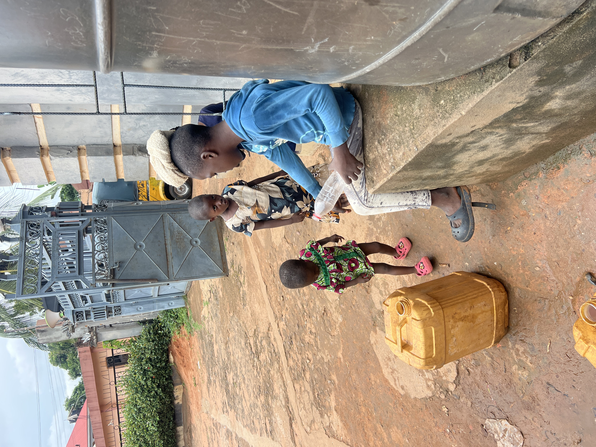 Three children are outdoors in Obosi, Nigeria, near a large gray water tank situated on top of concrete blocks. The ground is dry and cracked reddish-brown soil. One child in a blue long-sleeved shirt and light-colored pants pours water from a plastic bottle. Another child in a floral-patterned shirt looks on. A third child in a brightly patterned dress lies on the ground, next to a yellow jerrycan. A metal gate and fence are visible in the background.