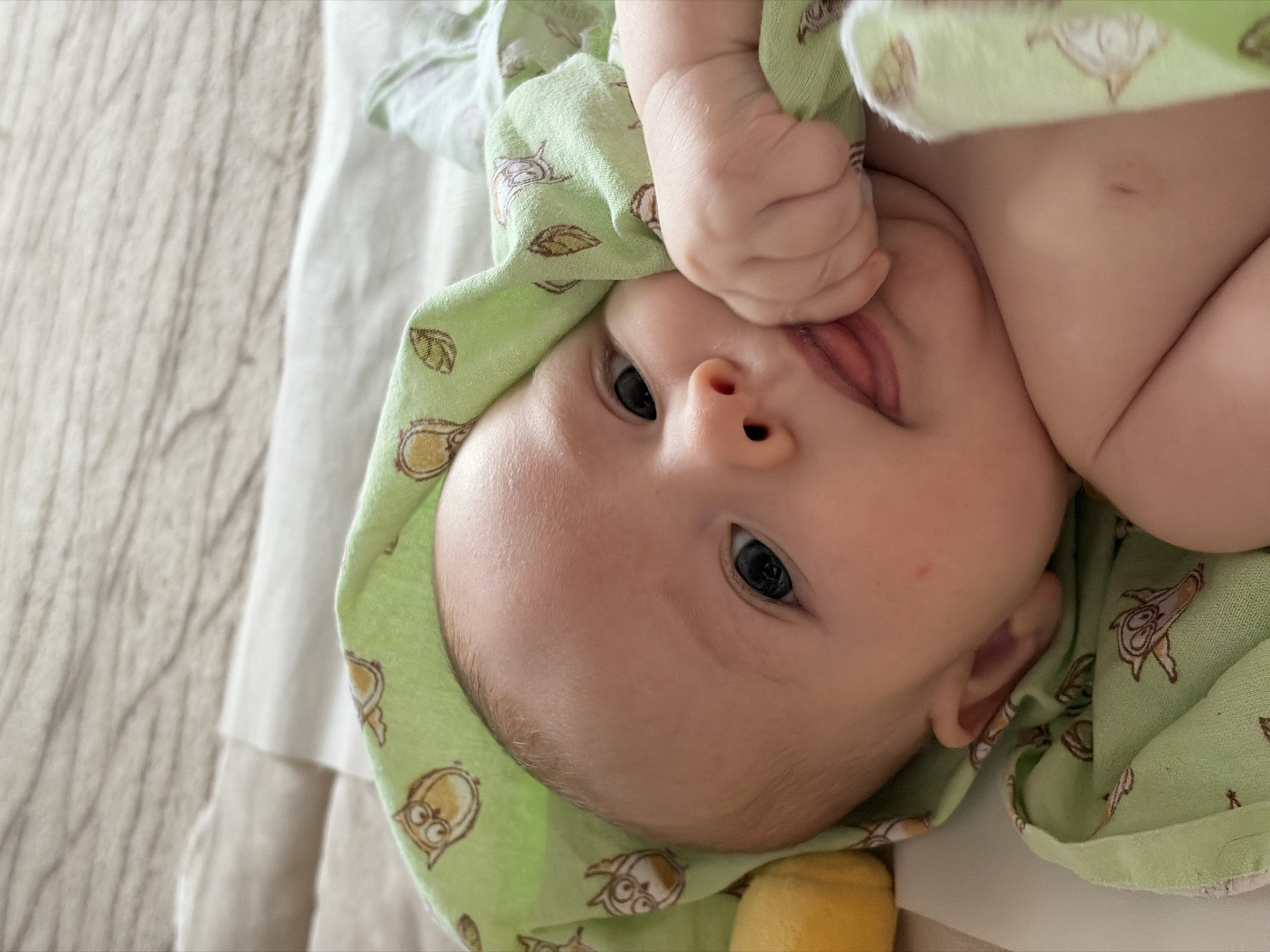 A baby lies on its back, partially wrapped in a light green cloth with a pattern of owls and leaves. The baby is fair-skinned and has dark blue eyes. One of its hands is raised near its face, with a finger touching its mouth. The background is blurred and appears to be a light-colored surface, possibly a bed or changing table.
There is no information in the image that indicates it was taken in Chaikovsky, Russia.