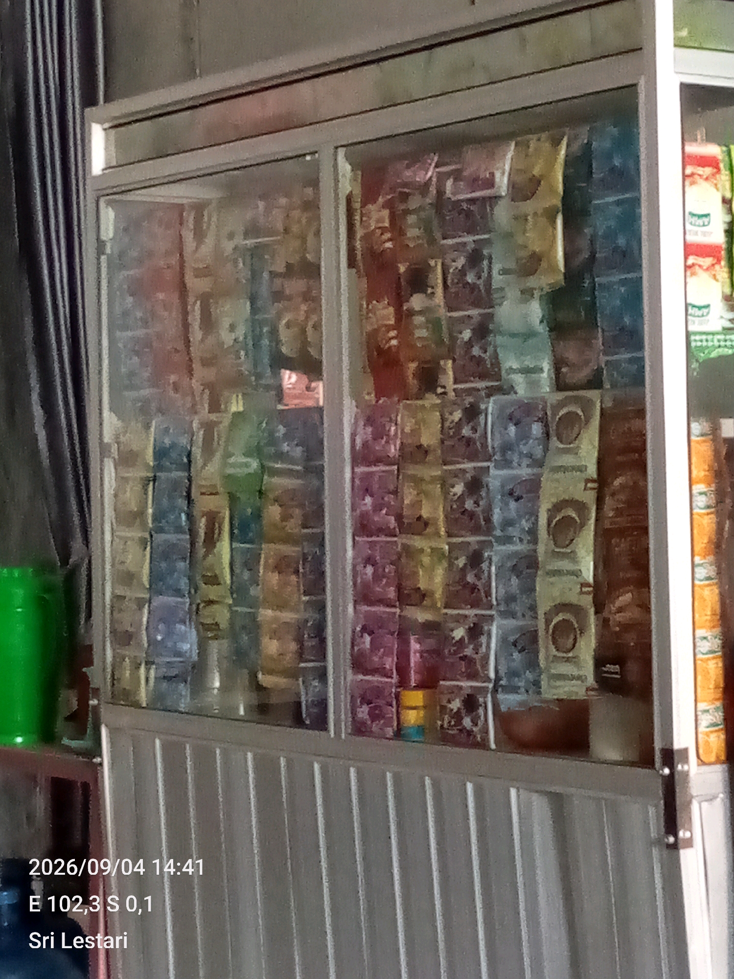 An outdoor photo captures a display shelf filled with various packaged goods, likely snacks or instant food items, organized in vertical rows behind a glass enclosure. The items are predominantly colorful, with red, yellow, blue, and green packaging visible. To the left of the display, a bright green container is partially visible, and a dark curtain or drape hangs to the far left. The bottom of the image shows text overlay with a date and time "2026/09/04 14:41", coordinates "E 102,3 S 0,1", and a name "Sri Lestari". The overall scene appears to be a storefront or market stall in Bukit Lembah Subur, Indonesia, with the display designed to attract customers.