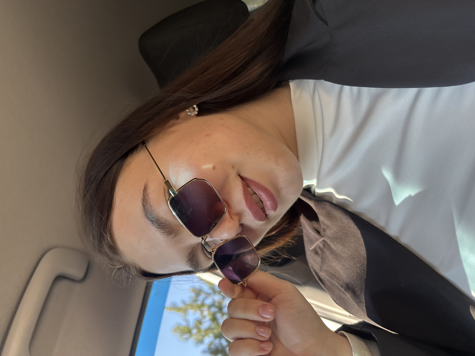 A woman with long brown hair is angled sideways in what appears to be the interior of a car. She is wearing sunglasses, a white top, and a black blazer. Her left hand holds the sunglasses near her face. A portion of a blue sky and tree canopies are visible outside the car window. The location is not discernible as Zaosinovo, Russia based on this image alone.