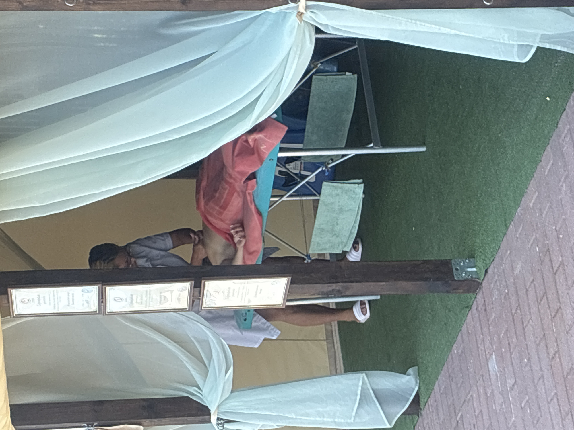 A person is receiving a massage on a massage table in an outdoor tent. The tent has sheer white curtains and is set up on a green turf surface, next to a brick walkway. A few framed certificates are hanging on a wooden post inside the tent. The person on the table is covered with a red towel and is lying on their stomach. The masseuse, wearing a blue shirt, is standing and appears to be working on the person's back. There are several green towels draped over the legs of the massage table. The setting appears to be a spa or wellness area, possibly at a resort or hotel in Sochi, Russia, given the context.