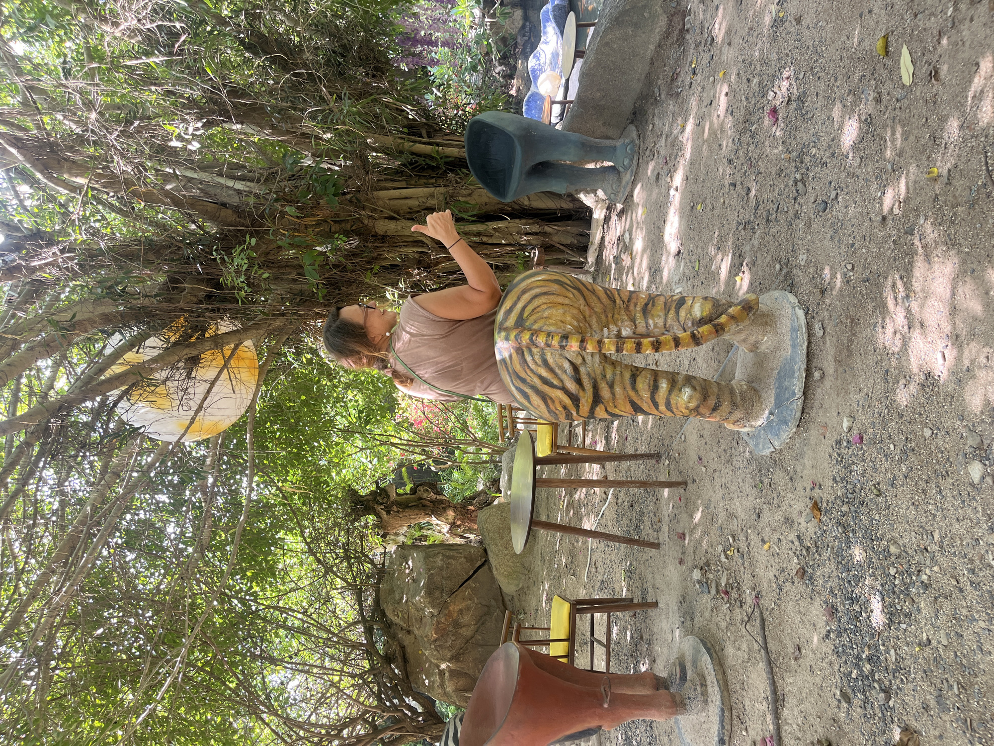 A woman with brown hair and glasses, wearing a tan shirt, stands in a garden and gives a thumbs-up. She is positioned behind a sculpture of tiger hindquarters. To her left is a round table with two yellow chairs, and to her right are two dark-colored sculptures resembling human legs in a seated position. The garden features a large tree with hanging vines, a large rock, and various plants and flowers. The ground is covered with dirt and gravel. In the background, a body of water can be seen. The scene appears to be an outdoor art installation or park located in Nam Nha Trang Ward, Vietnam.