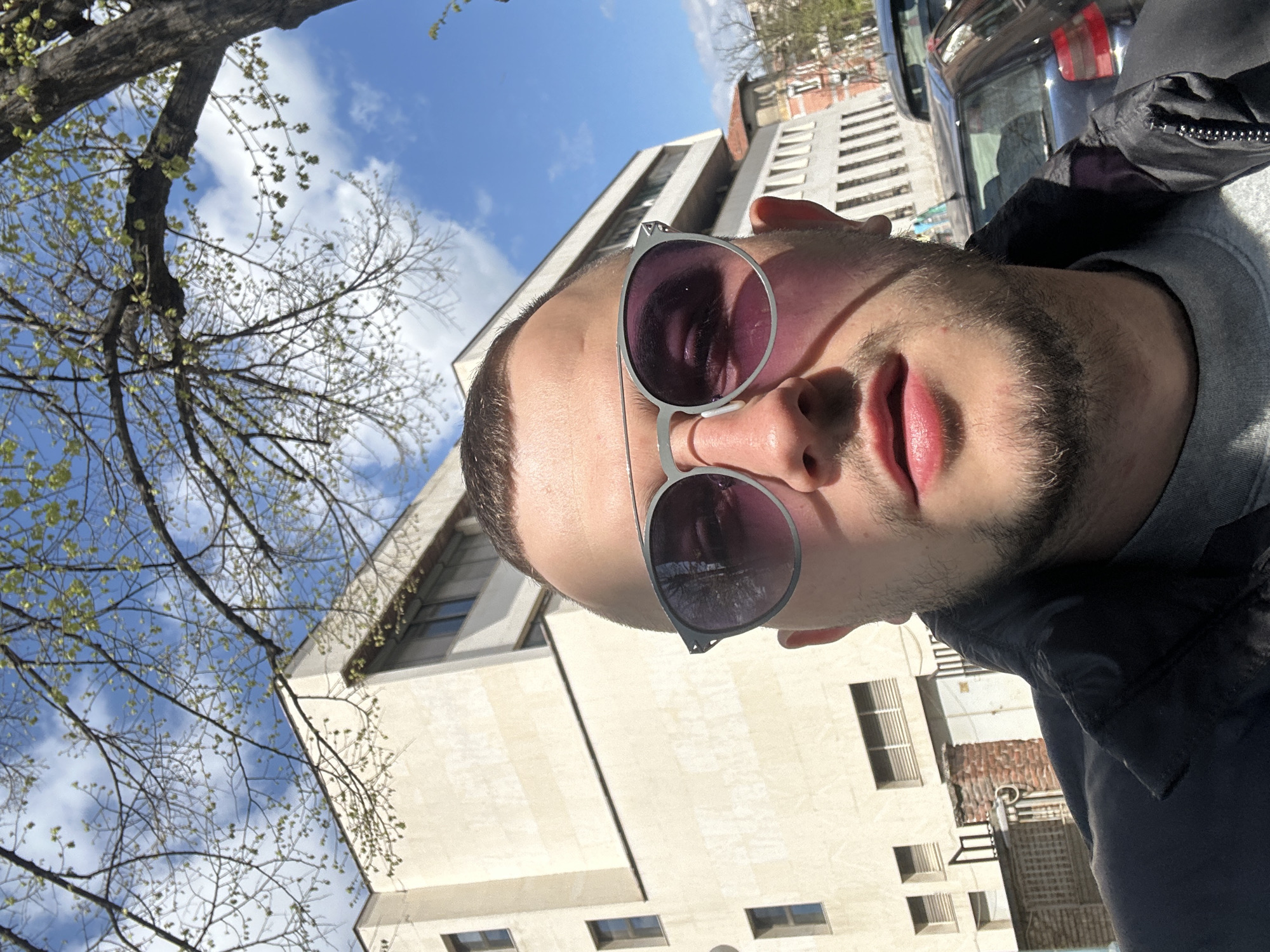 A low-angle, outdoor selfie depicts a man with short, dark hair and a beard wearing round sunglasses. A light-colored building with rectangular windows and a flat roof is behind him. A tree with budding leaves is to the left, against a blue sky with scattered clouds. A partial view of a parked car is visible on the right. Based on architectural style and available geolocation data, the building appears to be located in Plovdiv, Bulgaria.