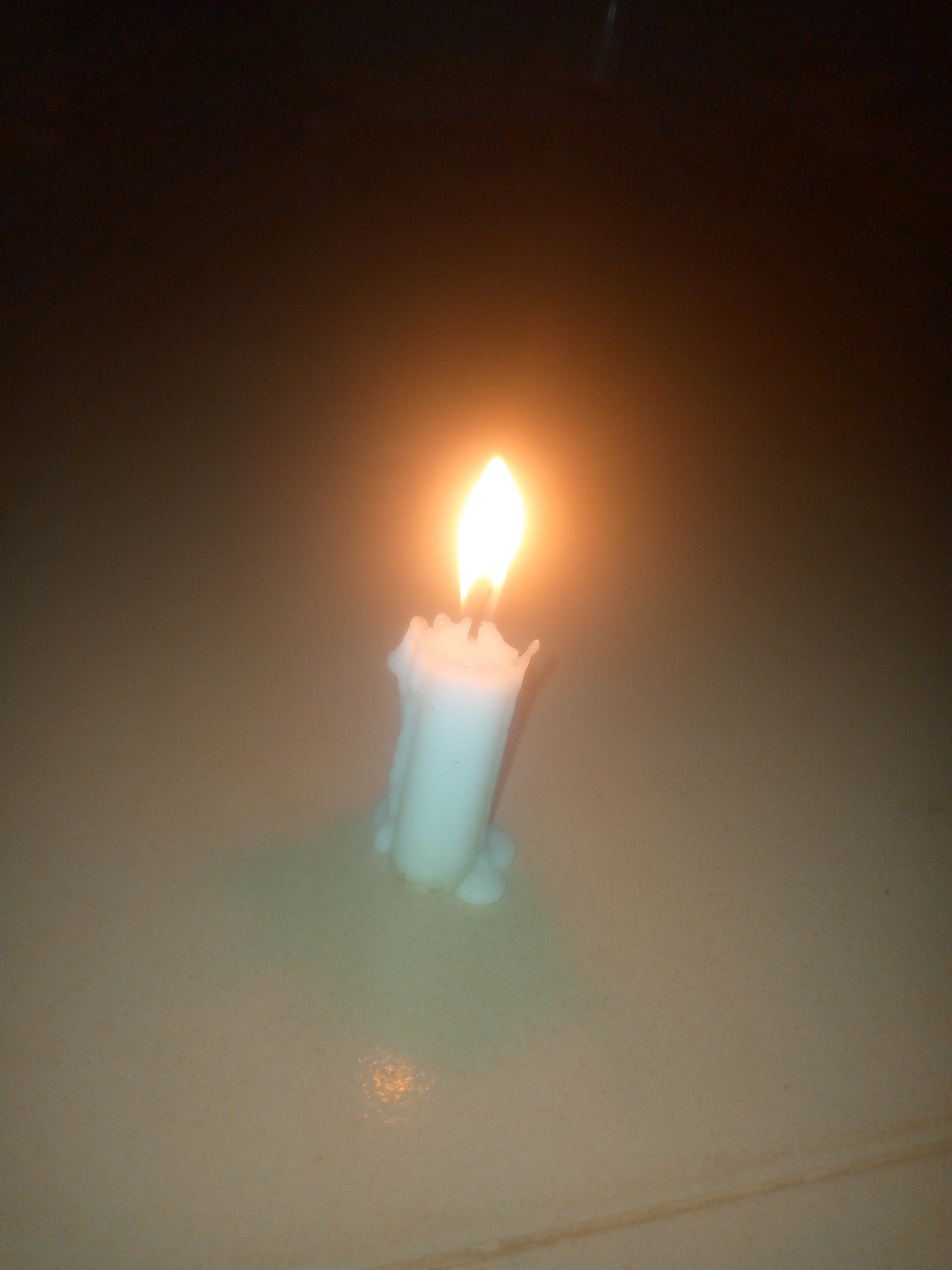 A lit, white wax candle sits on a tan tile surface in a darkened environment. The candle is partially melted, with wax drippings visible. The tile appears to have a reflective surface, mirroring the candlelight. The background is dark and out of focus. Location: Unknown, Unknown.
