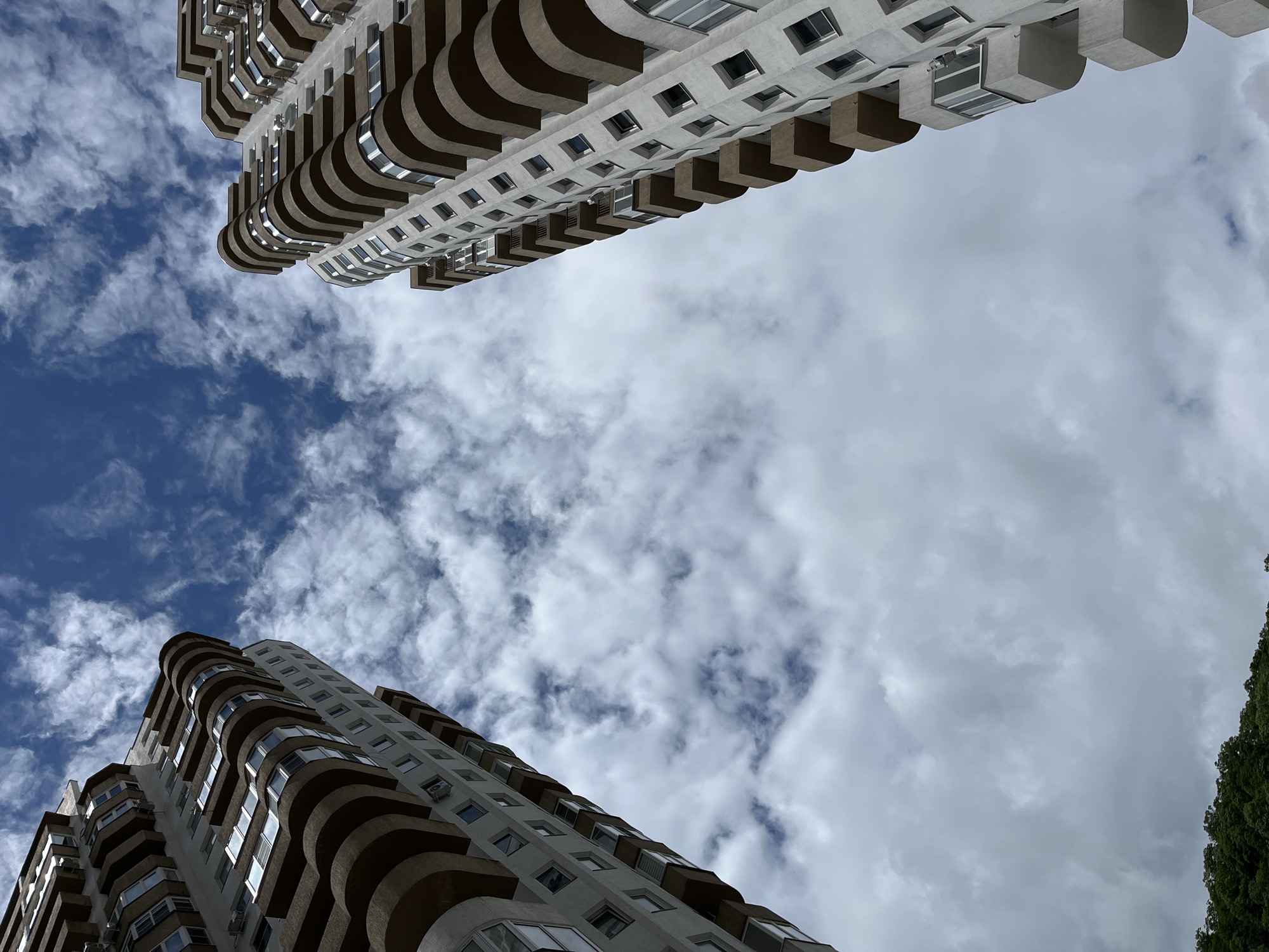 The image presents a low-angle shot of two tall buildings in Sochi, Russia, against a backdrop of a cloudy sky. The buildings, modern apartment complexes, are positioned at opposing corners of the frame. The building on the bottom left features curved balconies, and the one on the top right has squared balconies. The sky is predominantly white and grey clouds. The location in the city of Sochi is further suggested by the architecture.