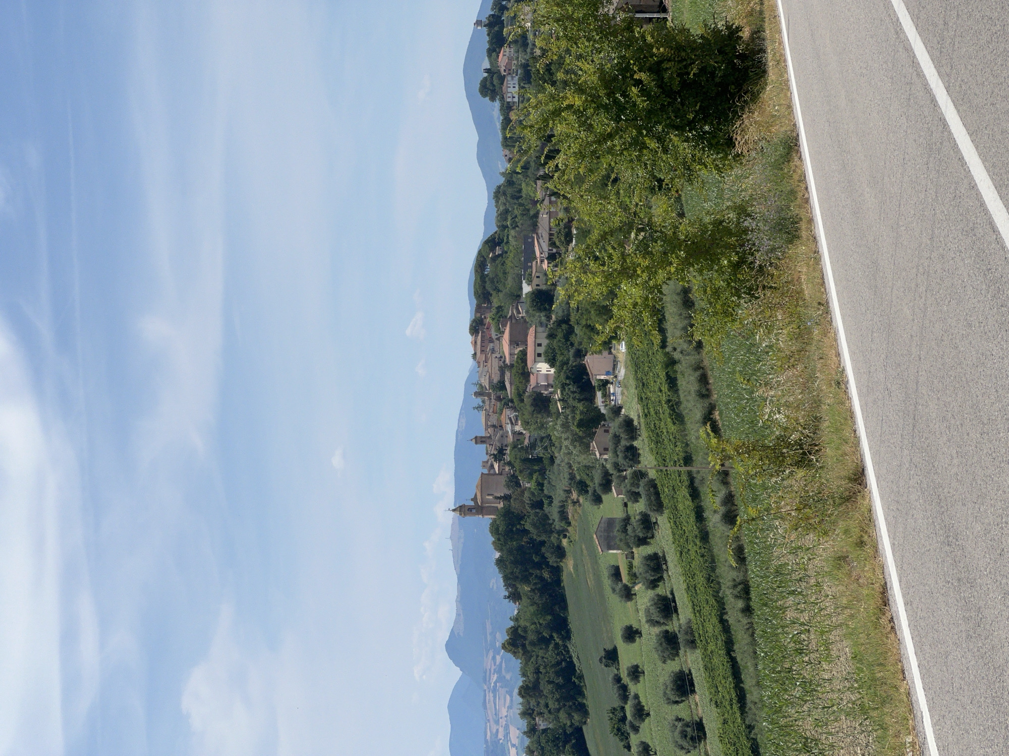 This is a full shot, eye-level, outdoor, daytime, landscape photograph taken from a road looking towards the Italian town of Barbara. The foreground consists of a paved road with a white line separating lanes, bordered by dry grass and some green vegetation. In the midground, there are rolling hills covered with agricultural fields. Some fields show rows of green crops, possibly corn, while others are planted with olive trees. A few small buildings are scattered among the fields. The background features the town of Barbara, situated on a hilltop. The town is characterized by terracotta-roofed buildings, with a prominent church tower. Lush green trees surround the town. In the far background, mountains are visible under a clear blue sky with scattered white clouds. The overall scene depicts a rural Italian landscape bathed in natural light.