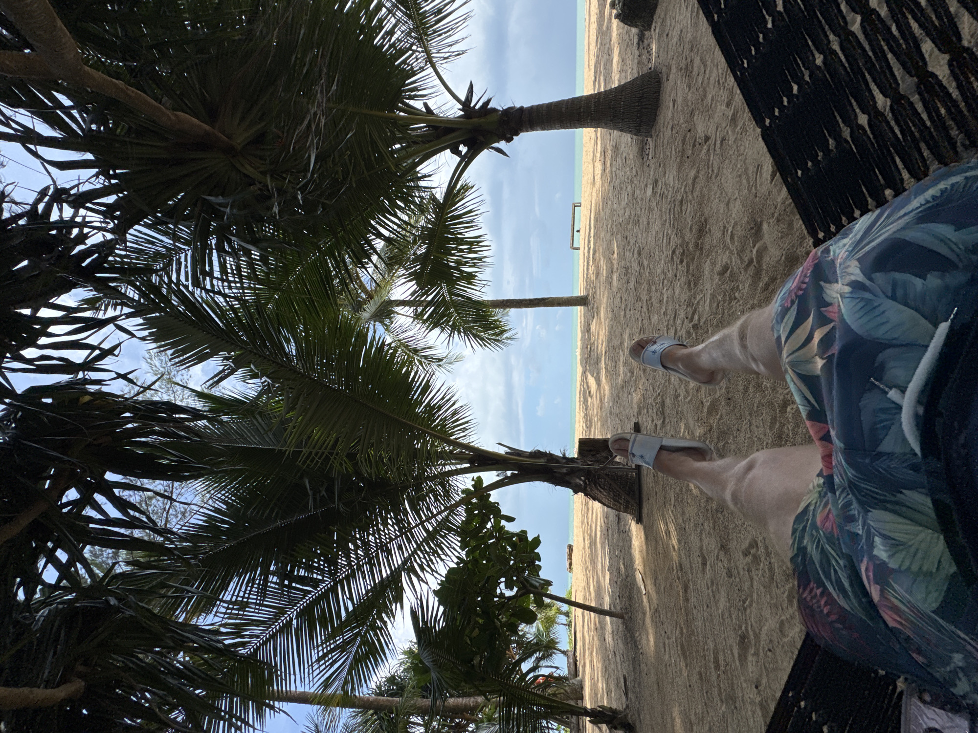 The image depicts a person's legs and lower torso, clad in floral-patterned shorts and light-colored slide sandals, lying in a black hammock. The hammock is positioned on a sandy beach, with the person's feet facing away from the viewer. The background reveals a tropical beach scene. Several palm trees with lush green fronds cast shadows on the sand, with their trunks visible. The sand is light brown and appears smooth, with some areas showing footprints or indentations from the hammock. Beyond the beach, a calm, turquoise body of water stretches to the horizon, meeting a cloudy sky with patches of blue. A small, dark-colored bench or structure is visible in the distance on the beach, near the water's edge. The overall environment suggests a relaxed vacation setting in Ban Khuk Khak, Thailand.