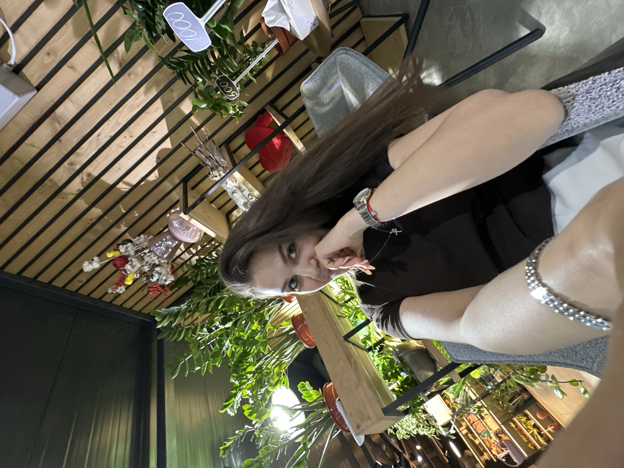 A young woman with long brown hair is posing for a selfie in an indoor setting, possibly a cafe or a retail space decorated with plants and wooden accents. She is wearing a dark brown t-shirt and a silver bracelet. Her right hand is touching her chin as she looks at the camera. In the background, there are several potted plants, shelves with decorative items, and two price tags with the numbers "600" and "1000" written on them. The overall atmosphere appears casual and relaxed.
