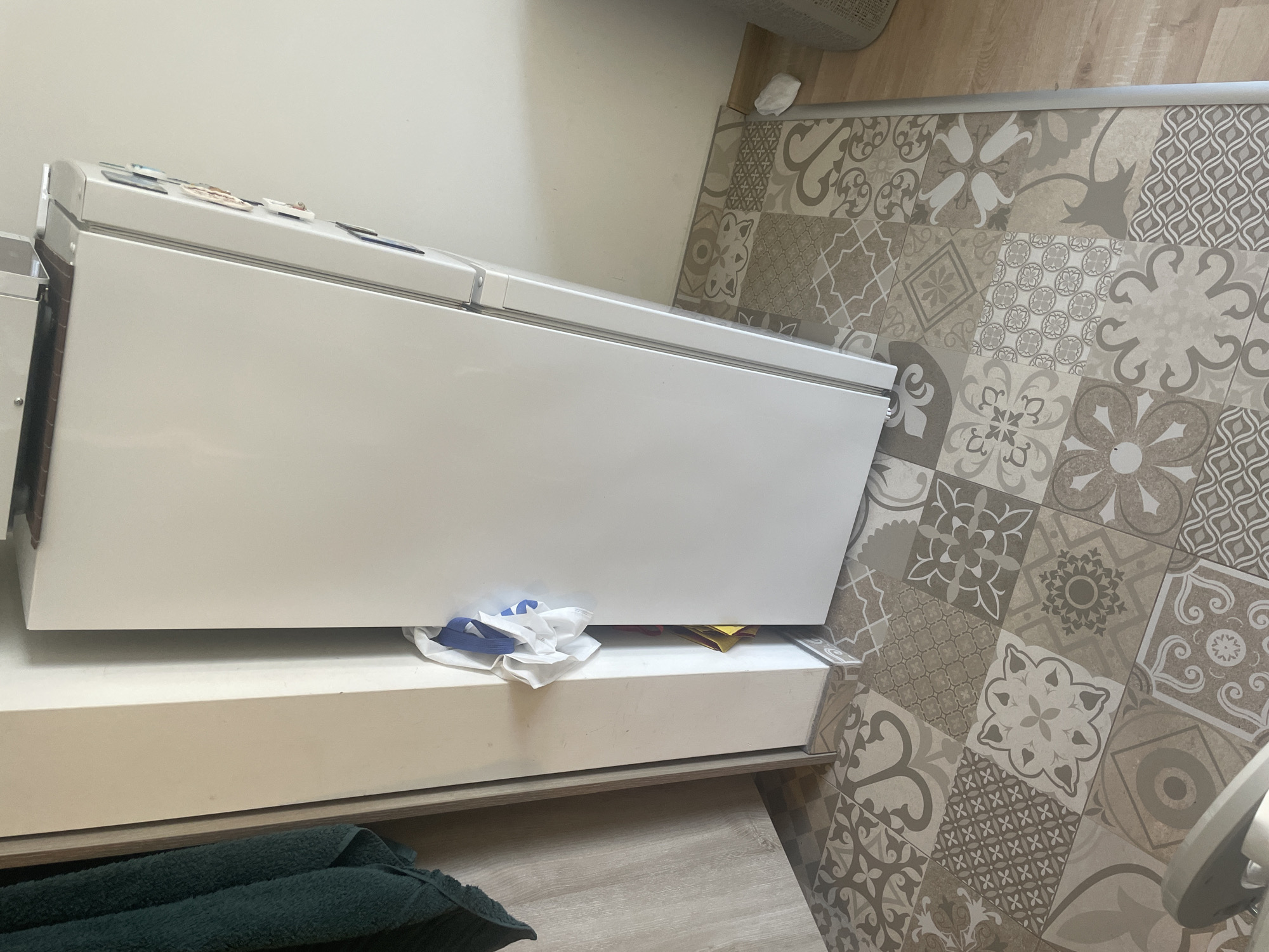 The image shows a corner of a room with a white refrigerator positioned on top of a white cabinet. The floor is covered with patterned tiles in beige and gray tones. A crumpled white cloth is visible between the cabinet and the refrigerator. Part of a dark green towel is visible in the lower left corner. The location is a room in a house, no information about the city of Sofia, Bulgaria is available. No people are visible in the image.