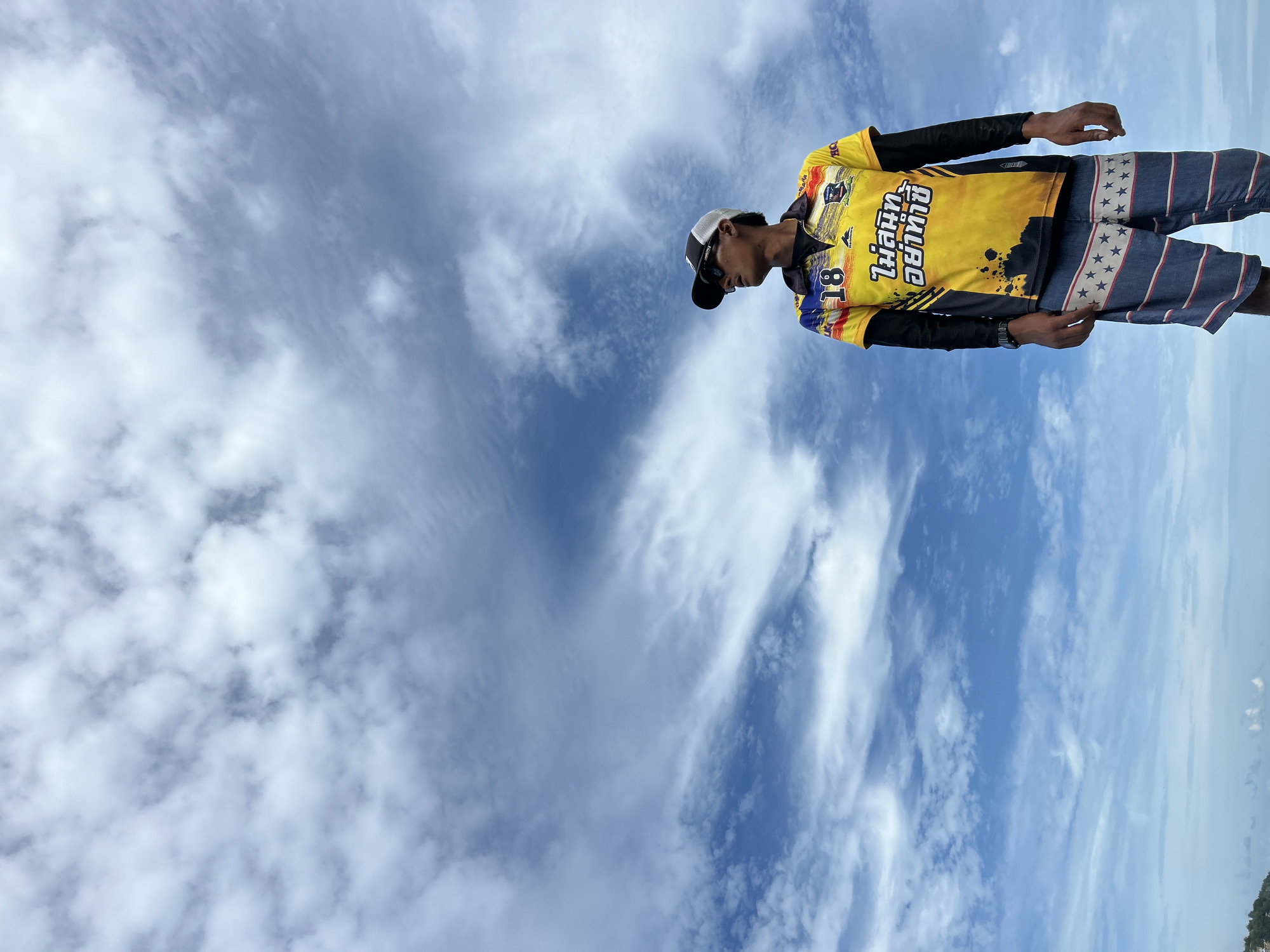 A young man stands outdoors, looking to his right. He wears a bright yellow short-sleeved shirt with a black collar, featuring colorful graphics and Thai text. Beneath this, he has a long-sleeved black shirt. His shorts are blue and white striped with a pattern of red stars. He also wears a baseball cap with a white front and a dark brim, and dark sunglasses. The background is a vast blue sky filled with dynamic, wispy white clouds. The lower part of the image shows a faint horizon line, suggesting a body of water, possibly the ocean, and a hint of green foliage to the left, indicating a coastal area. The setting is consistent with Ao Nang, Thailand, a popular beach destination known for its dramatic limestone cliffs and clear waters.