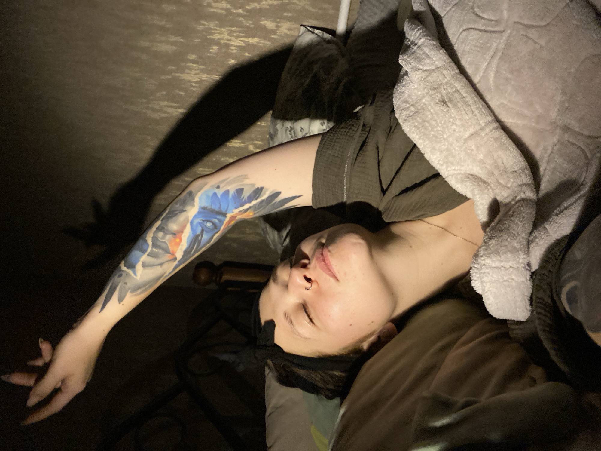 A person is lying in a bed, covered with a light-colored textured blanket, facing upwards. The person has a visible arm with a detailed tattoo, outstretched towards the left side of the frame, casting a shadow on a wall. The location appears to be indoors, likely a bedroom. The city of Chaikovsky, Russia is not discernible from the provided image.