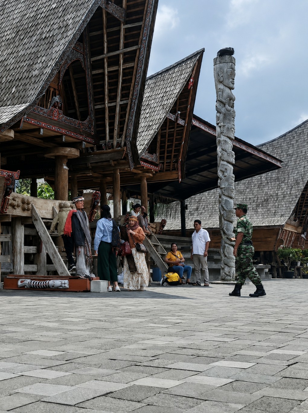 In Ambarita, Indonesia, a group of people are gathered in an open courtyard in front of traditional Batak architecture. The buildings feature distinctive, high-pitched roofs covered in wooden shingles and ornate, dark wood trim. A tall, carved stone totem stands prominently to the right of the frame, depicting a series of human faces stacked vertically.
In the foreground, the ground is paved with large, grey square tiles. Several individuals are interacting or observing the surroundings. On the left, a mannequin dressed in traditional attire stands near a carved wooden object. Two women are standing near the mannequin, one in a blue top and dark pants, the other in an orange headscarf and a patterned dress. Further back, a man in a white shirt and tan pants stands, and a woman in a yellow shirt sits with a yellow bag beside her. To the right, a soldier in camouflage uniform walks away from the camera, his back facing towards the viewer. The overall atmosphere appears to be a cultural or historical site.