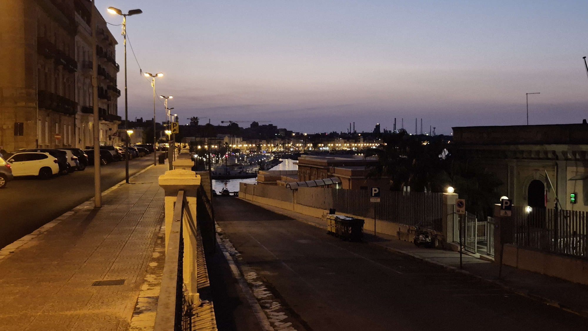 The image captures a dusk scene in Taranto, Italy, specifically along the Lungomare. On the left, a pedestrian walkway lines the street, with parked cars stretching into the distance. Buildings with illuminated windows and balconies rise on the left side of the street. The street itself is paved, with lampposts illuminating the sidewalk.
The scene then transitions to a view overlooking a harbor, with boats docked and lights reflecting on the water. In the background, the city skyline is visible, dotted with lights and industrial structures. On the right side of the street, a low wall separates the road from a pathway, beyond which is a fenced area. A small dumpster is situated on the road. Parking signs are visible along the street. The sky is a soft gradient of purple and orange, indicative of twilight.